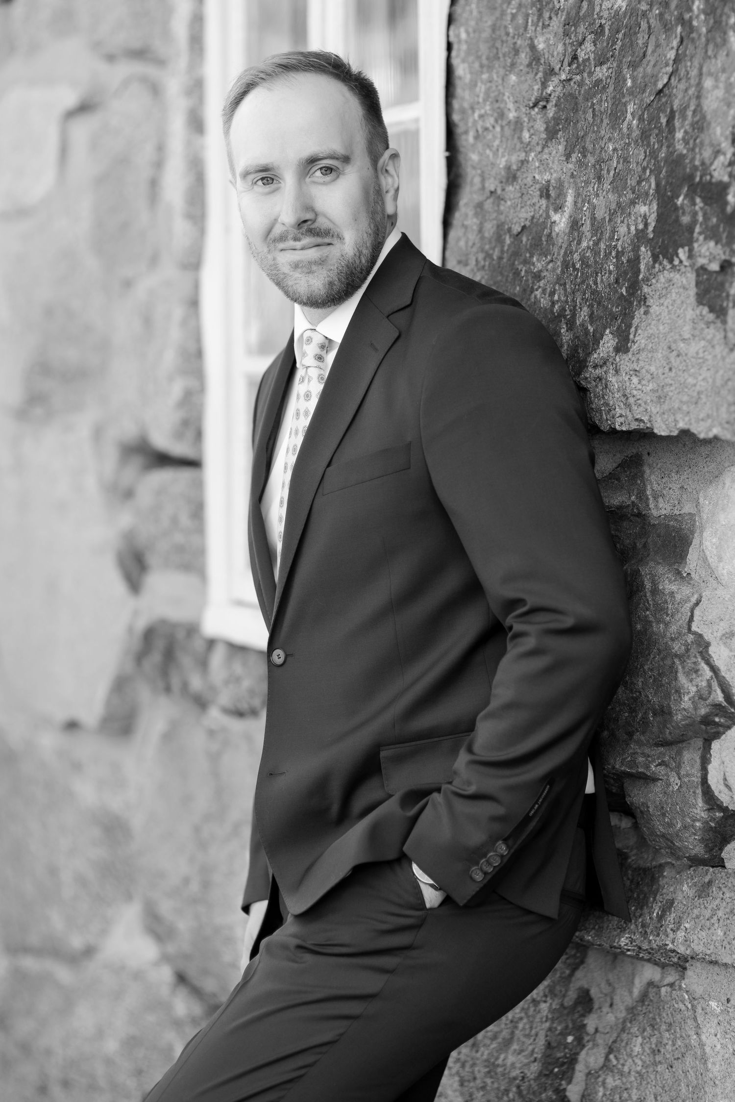 Casual portrait pose leaning against textured stone wall in formal business attire.