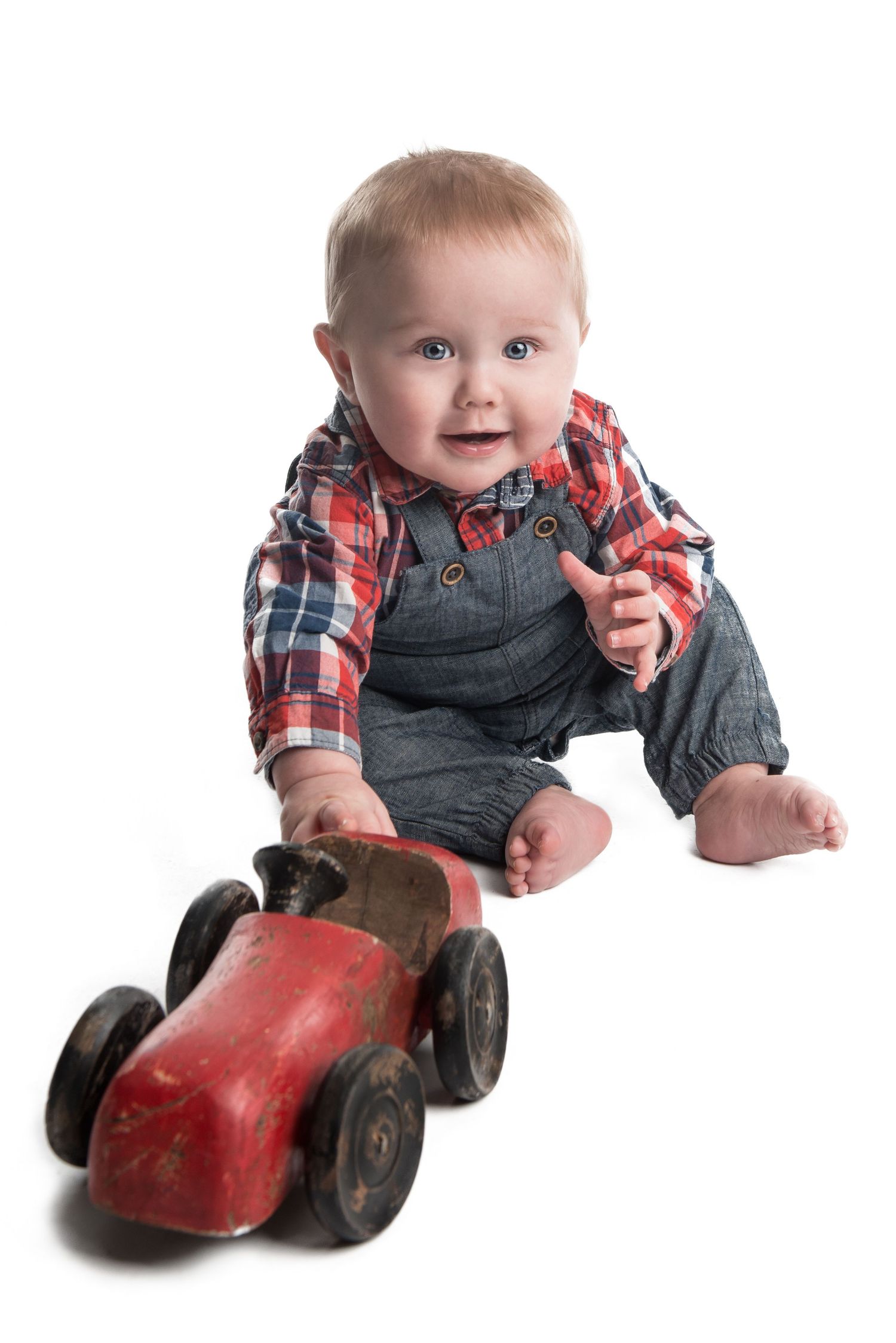 Baby in plaid shirt and overalls playing with vintage red toy car.
