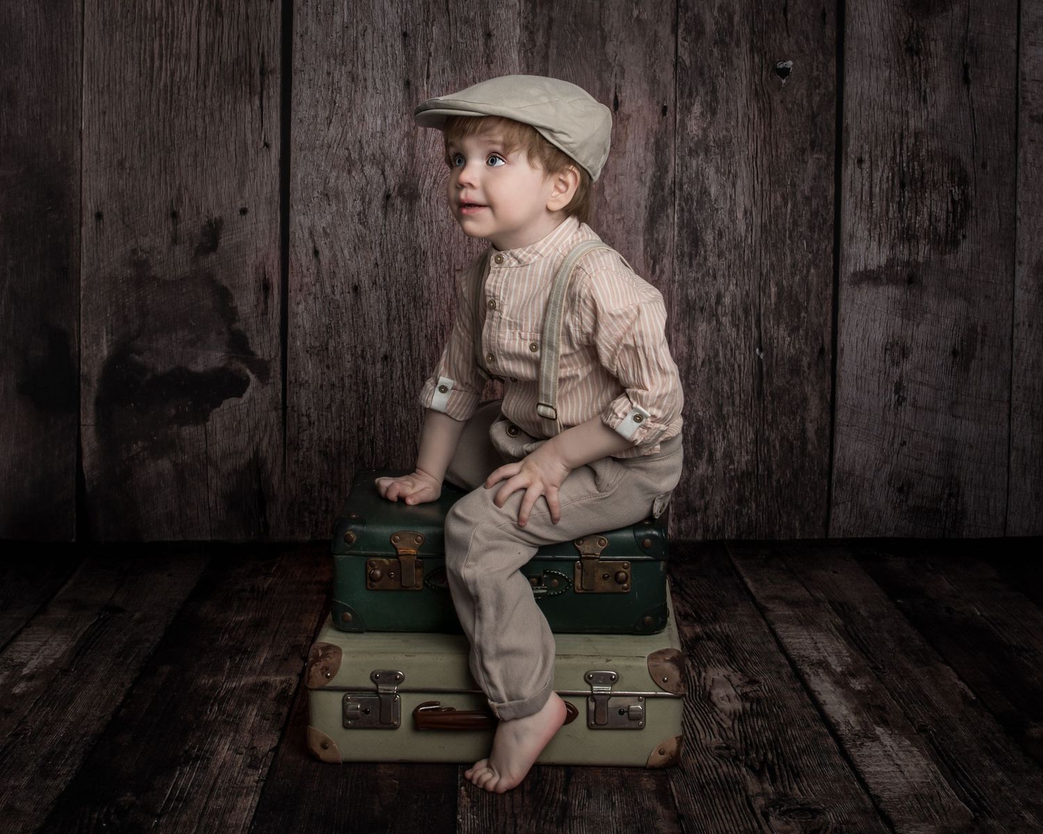 Vintage styled photo of a young child in newsboy cap and suspenders sitting on antique suitcases against wooden backdrop.