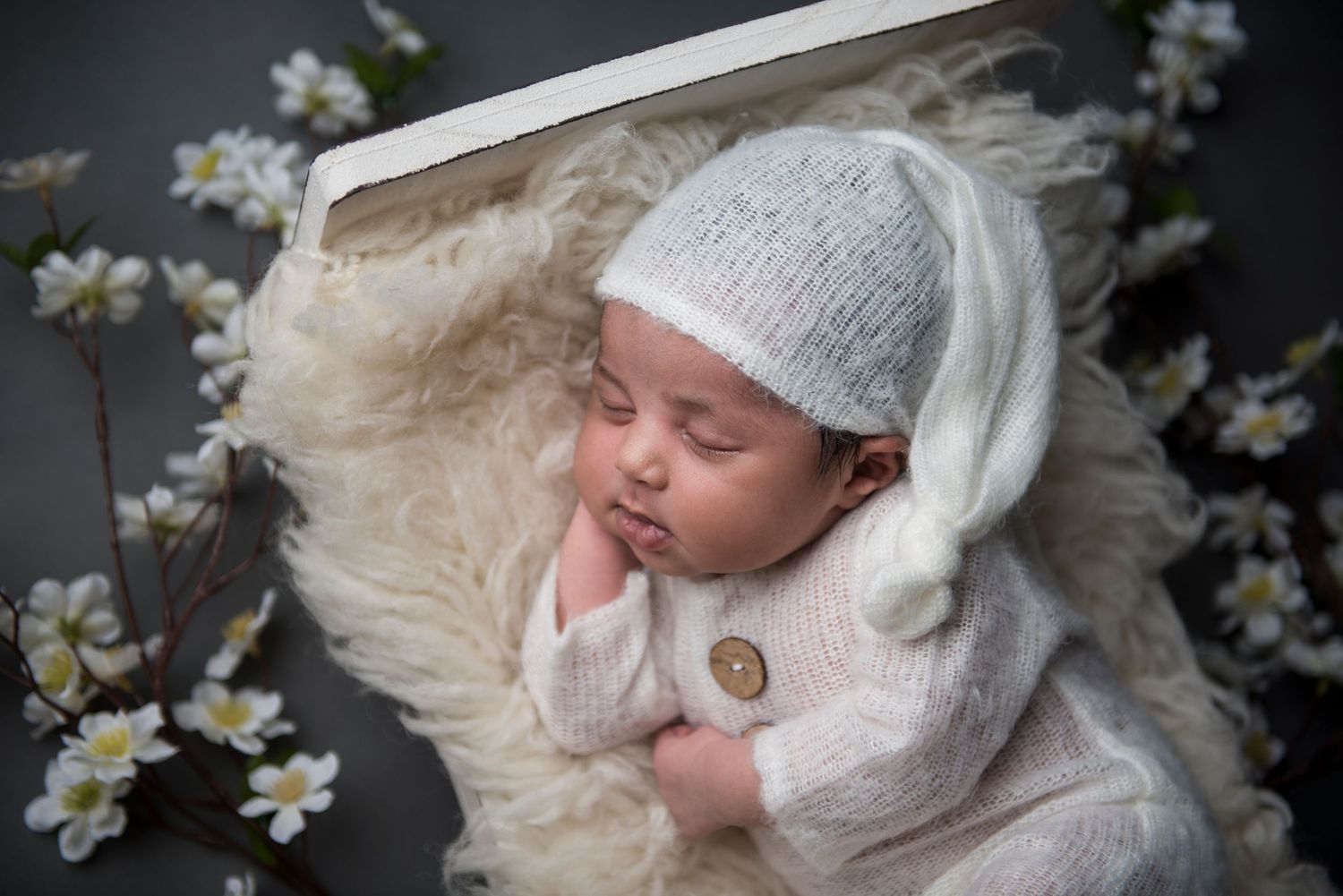 Sleeping newborn in knit outfit surrounded by white flowers and soft fur backdrop.