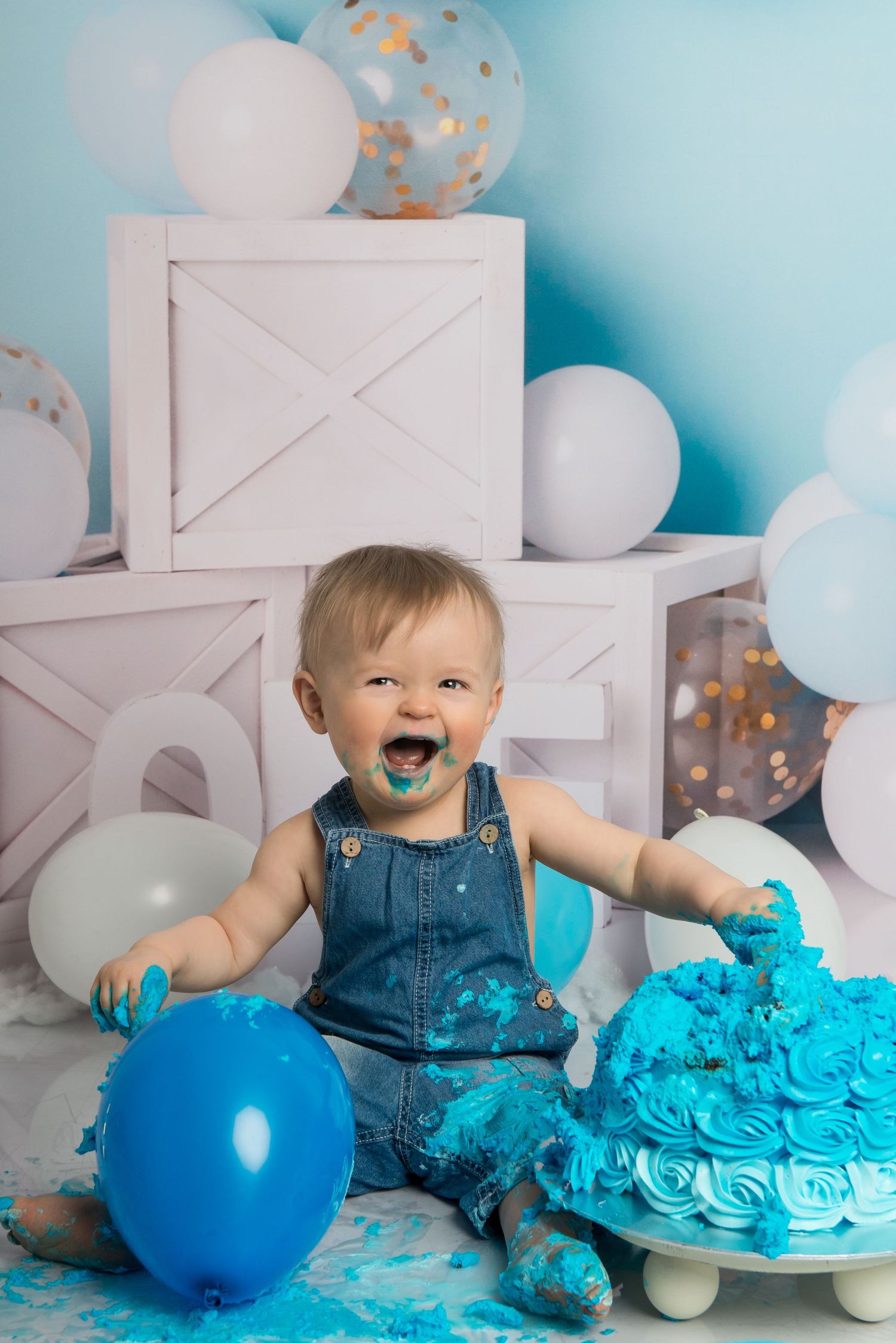 A festive first birthday cake smash with blue frosting, balloons, and denim overalls creating a messy celebration.