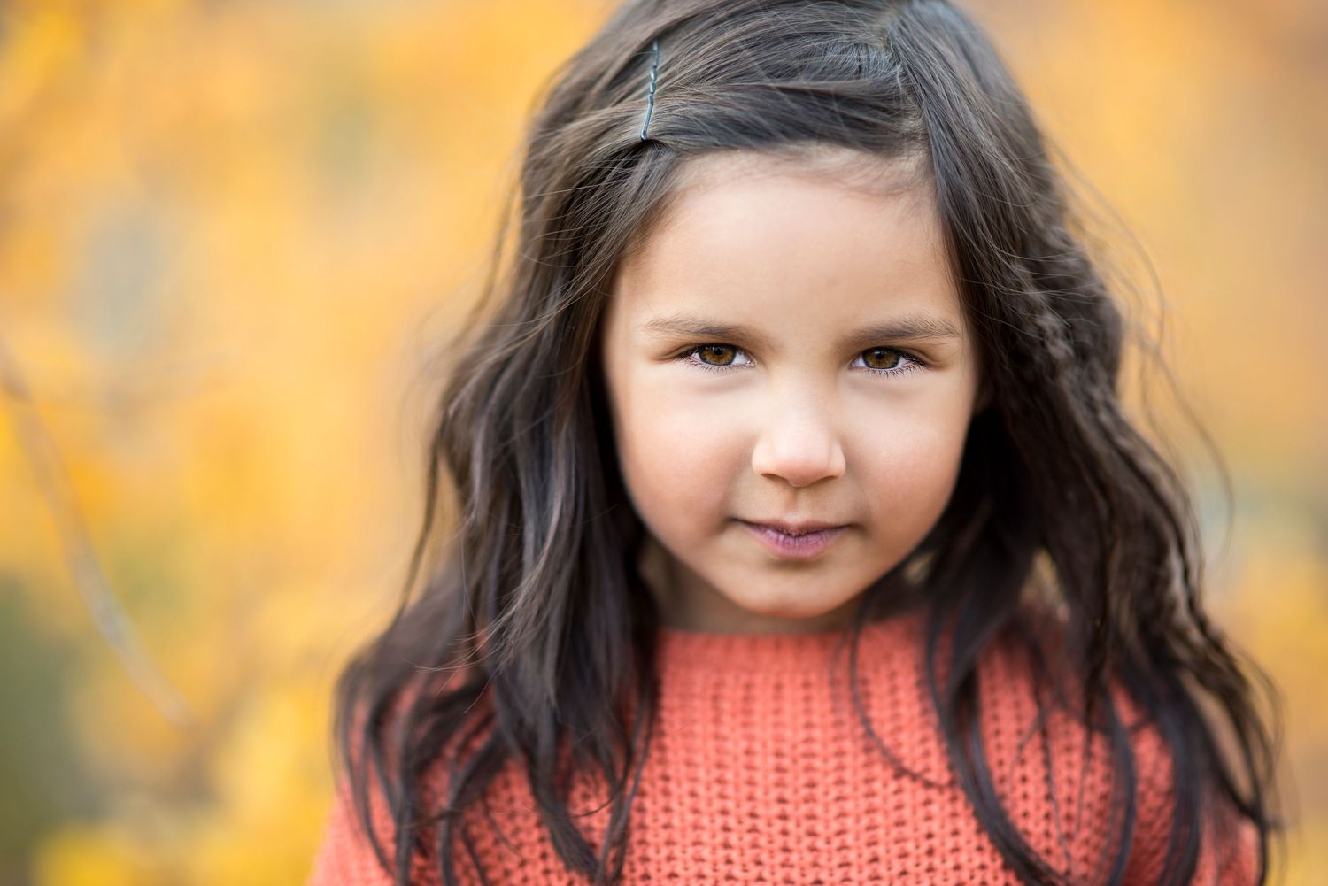 Young girl with long dark hair wearing a coral knit sweater, posing outdoors with golden autumn foliage background.