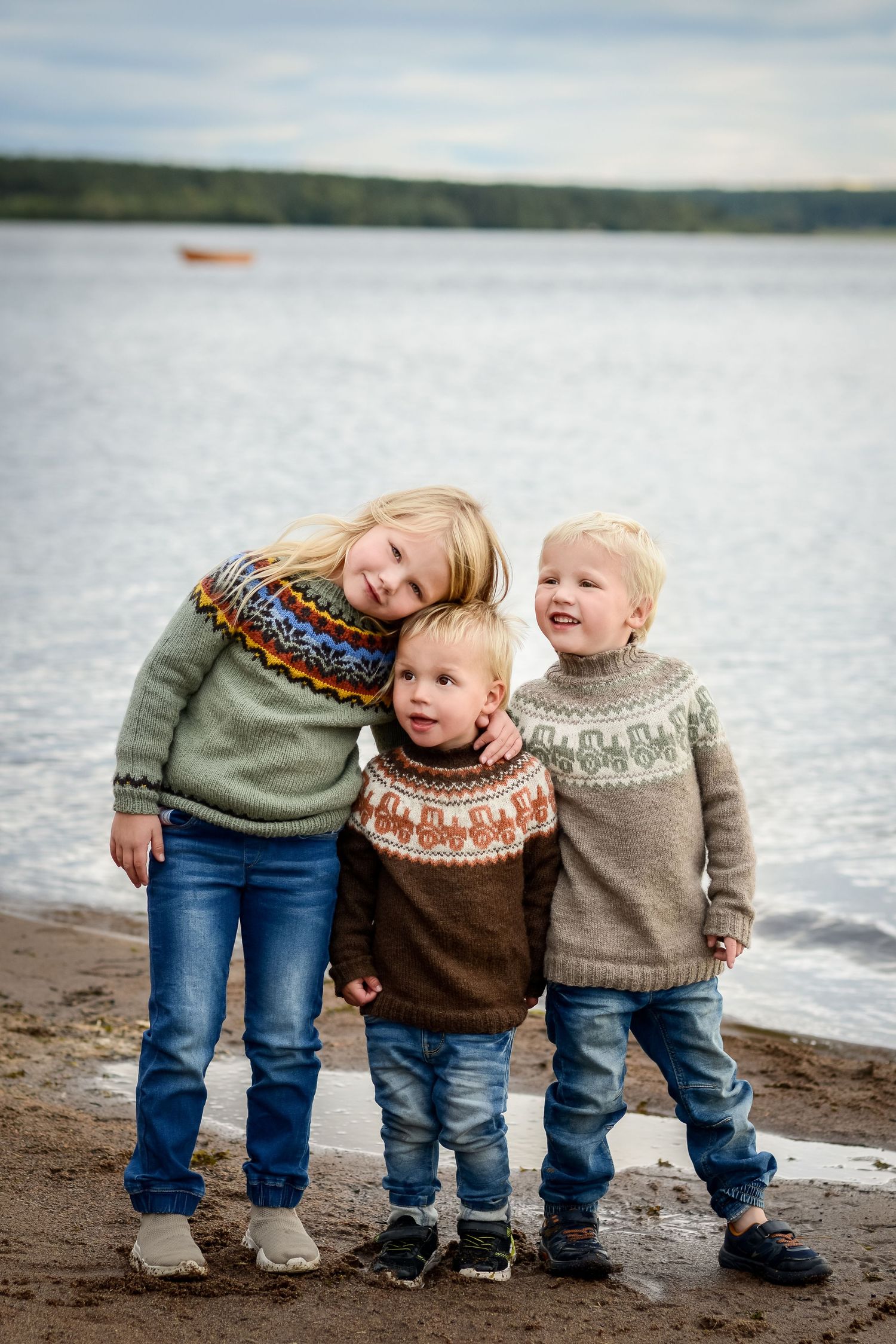 Three young siblings in colorful Nordic sweaters posing together on a sandy lakeside beach.