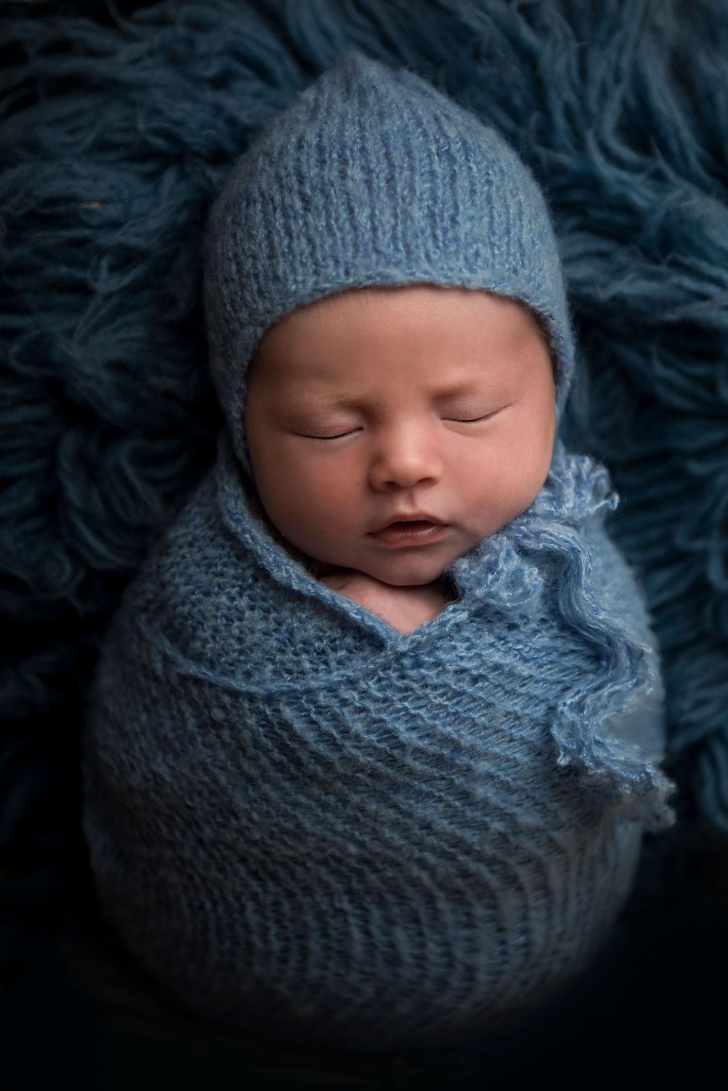 Sleeping newborn wrapped in blue knit cocoon against dark textured background.