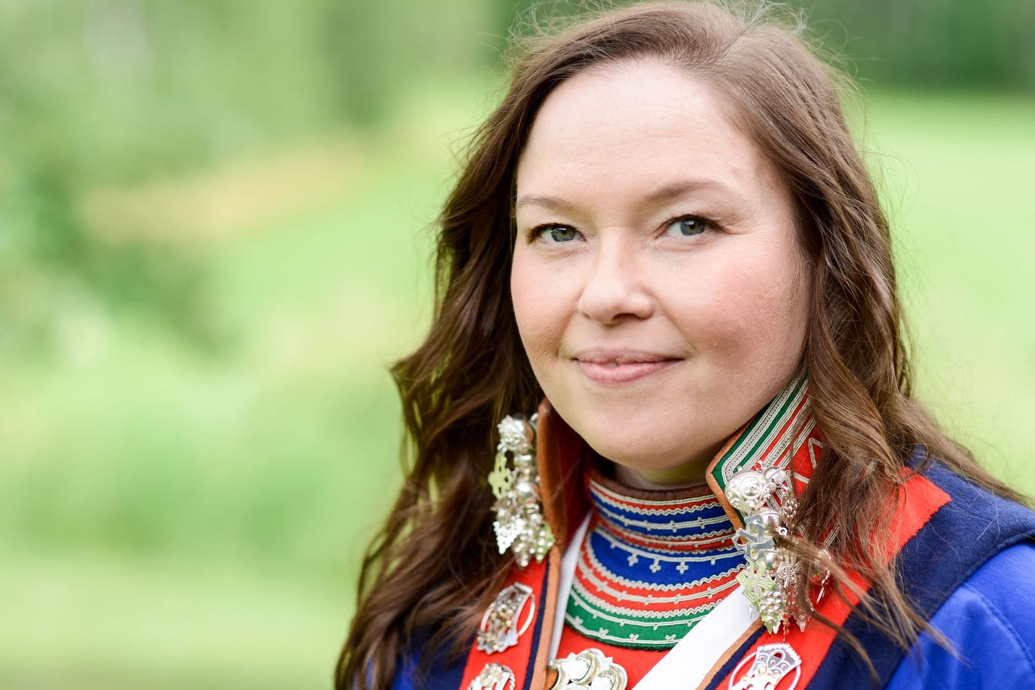Woman in traditional Sami cultural attire with colorful embroidery and silver jewelry, smiling outdoors.