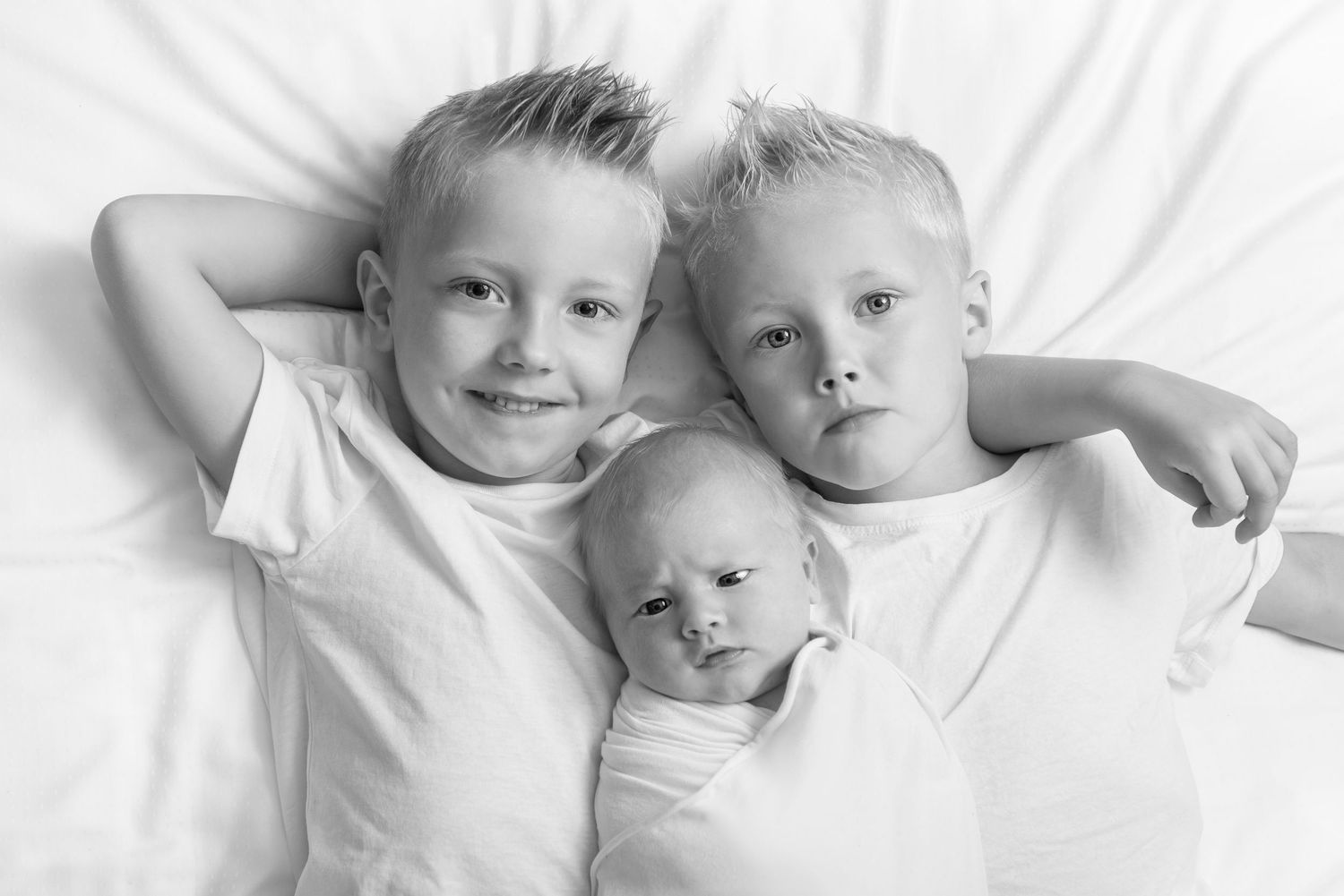 Three siblings in white clothing laying together on white bedding.