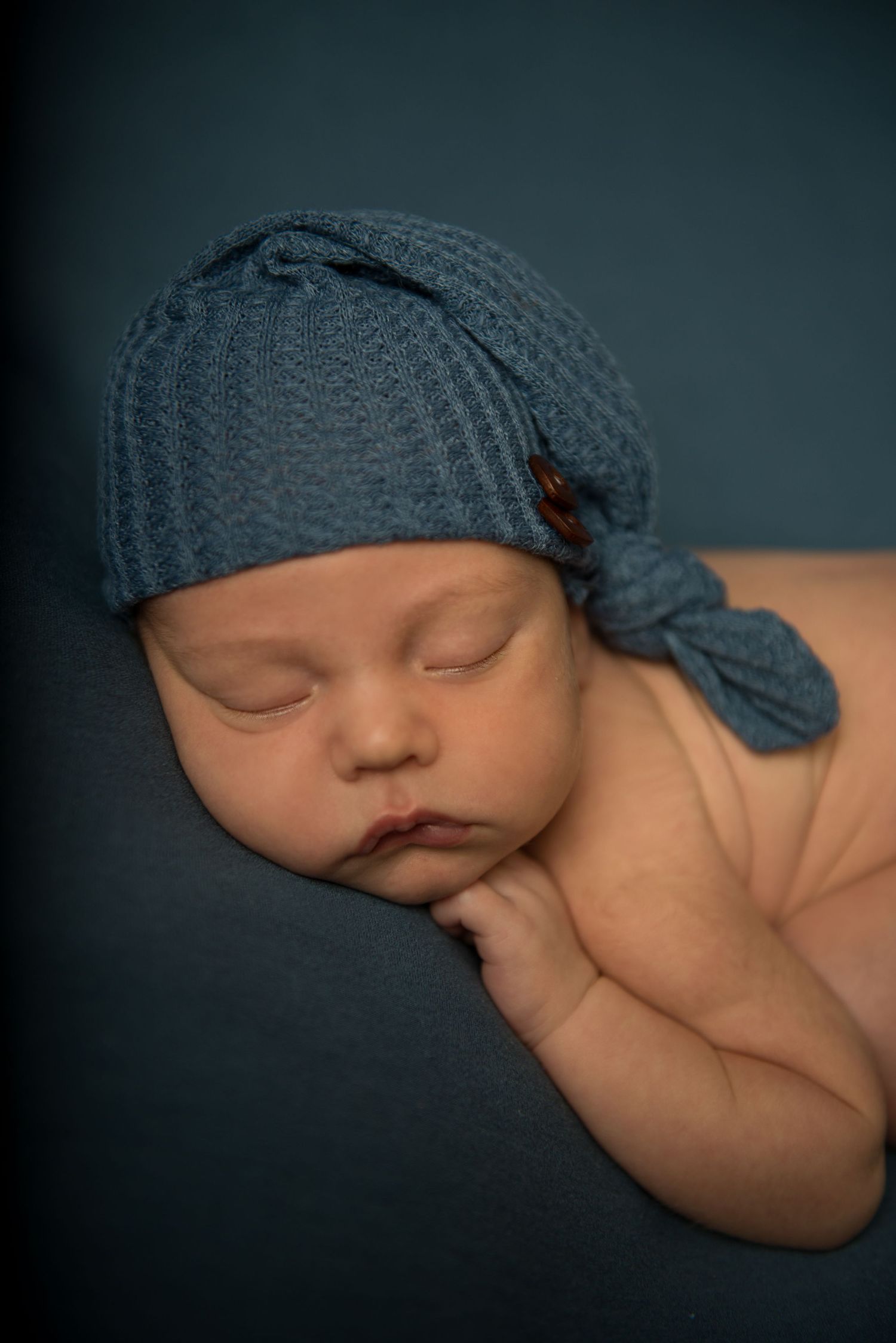 Peaceful sleeping baby wearing blue knotted cap against dark background.