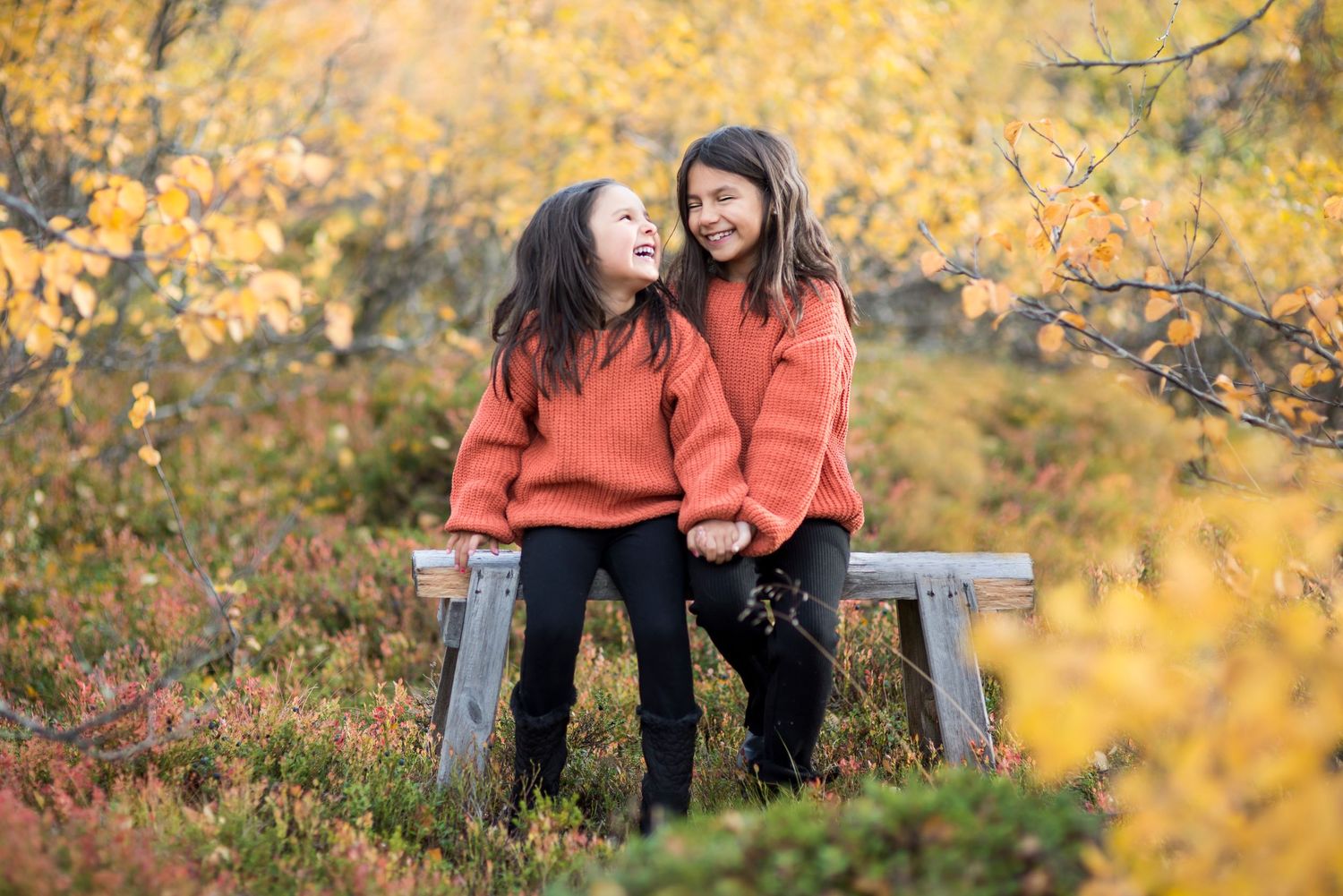 Two girls in matching red sweaters sit on a bench laughing together in an autumn forest setting.