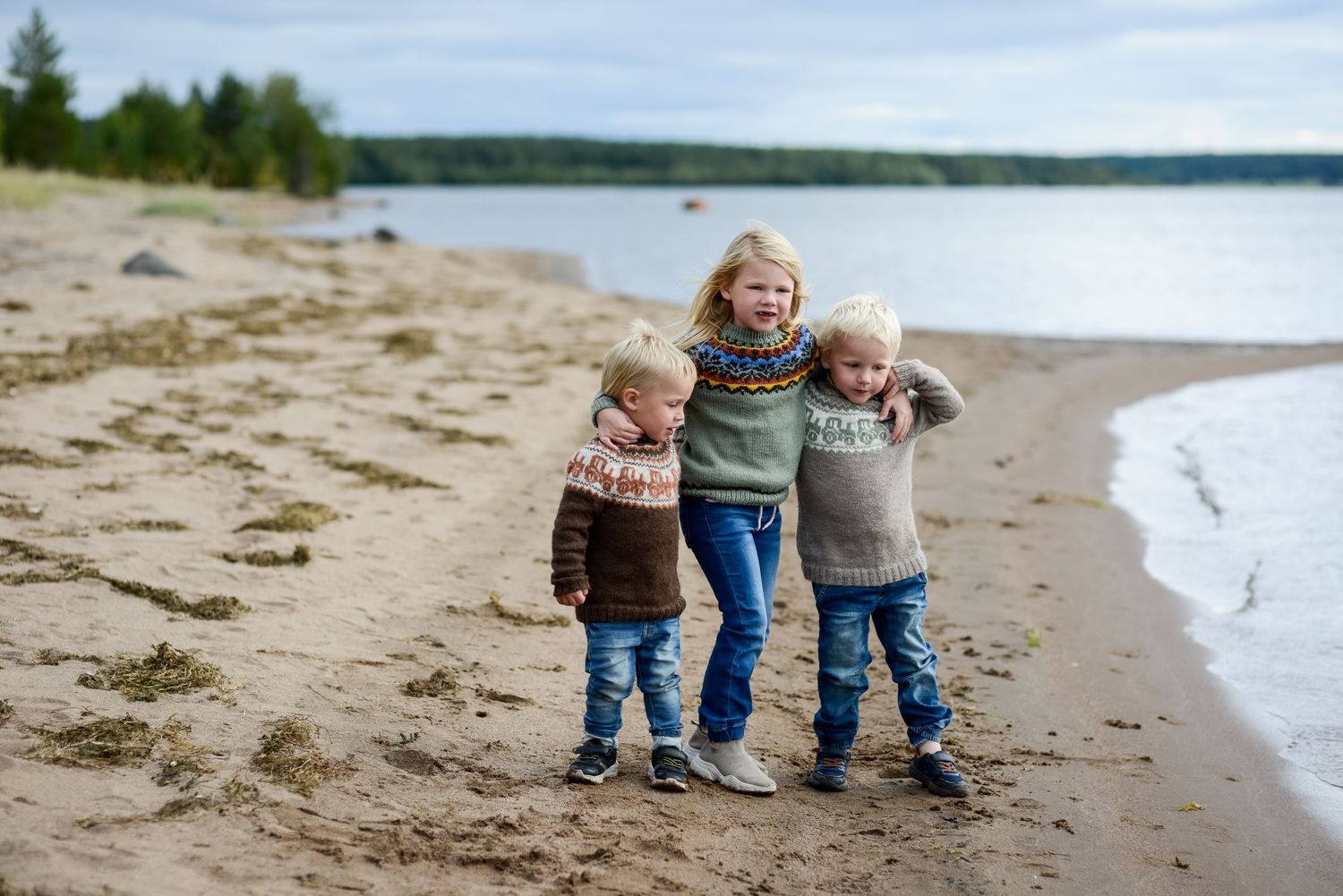 Three children in Nordic sweaters hugging and smiling on a sandy beach shoreline on a cloudy day.