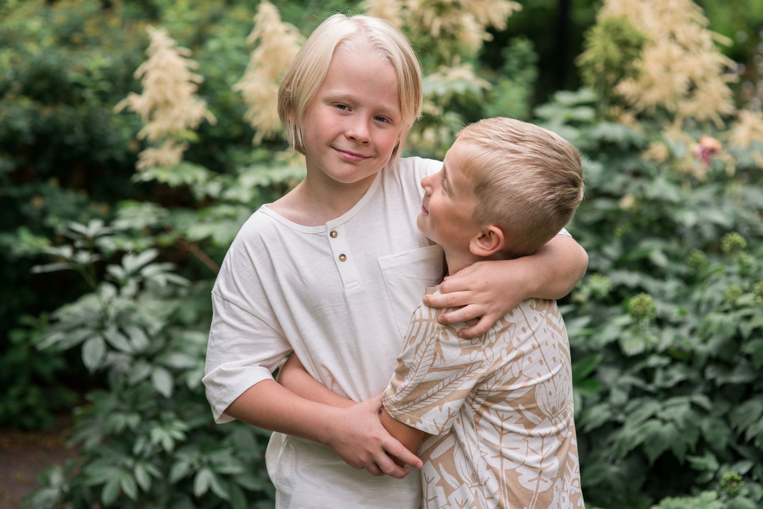 Two blonde children hugging outdoors in a lush green garden, smiling warmly in casual summer clothing.
