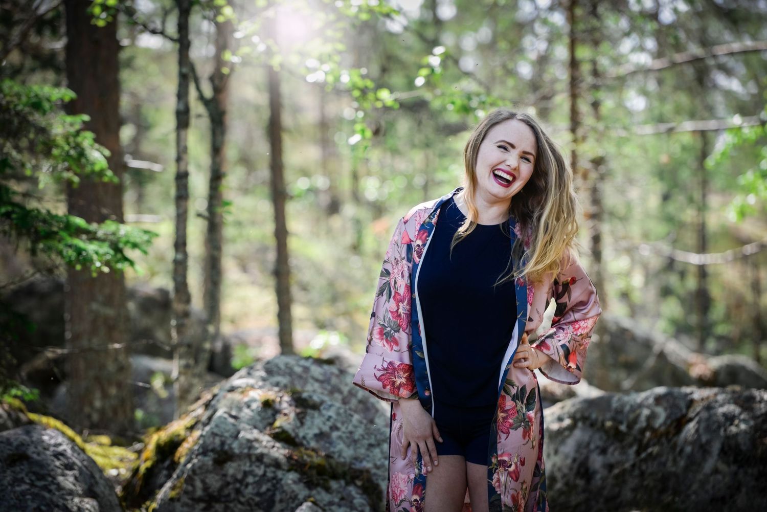 A laughing blonde woman in a floral jacket poses among mossy rocks in a sunlit forest.