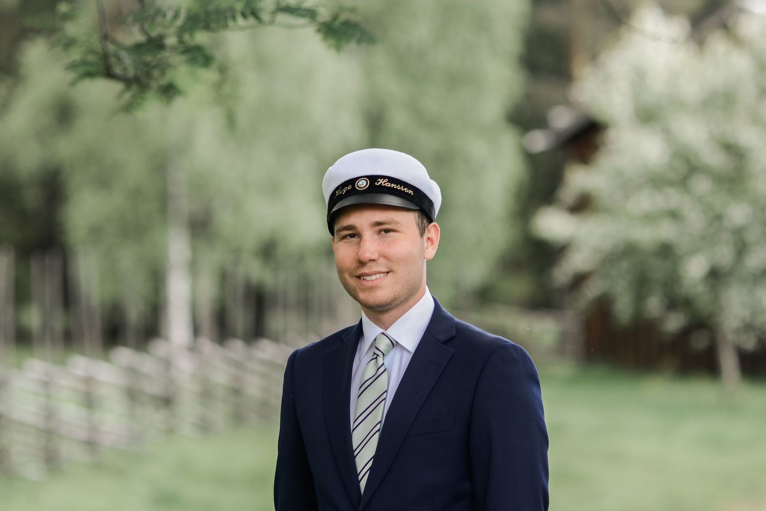 A person wearing a navy suit and cap poses in a serene outdoor garden setting.