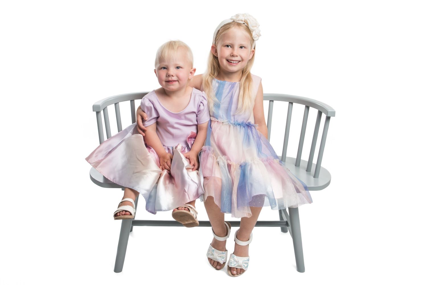 Young children posed on gray bench in matching pink and blue dresses.