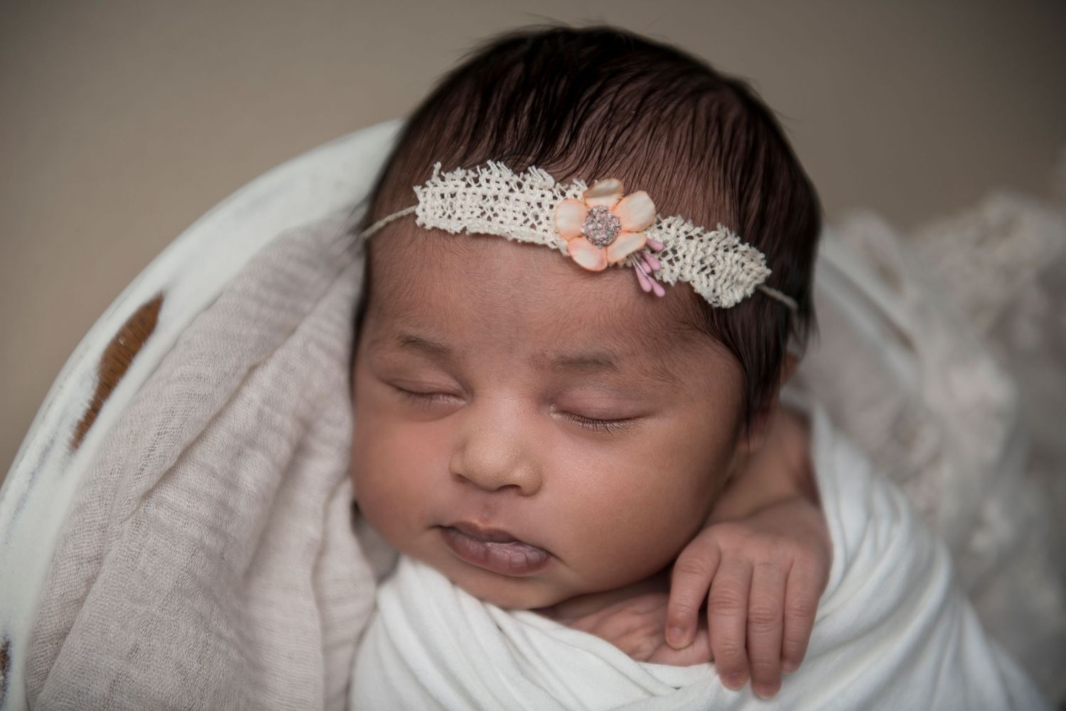 Sleeping newborn swaddled in white with delicate lace and pink floral headband resting on soft fabric.