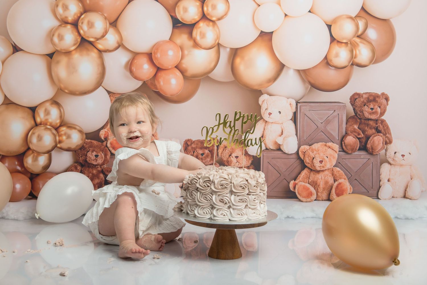 A baby reaches for a beautifully decorated cake against a balloon and teddy bear backdrop.