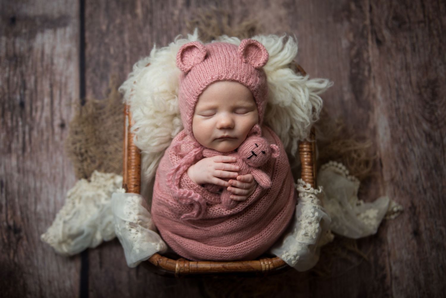 Sleeping infant wrapped in pink swaddle on rustic wooden backdrop with white lace accents.