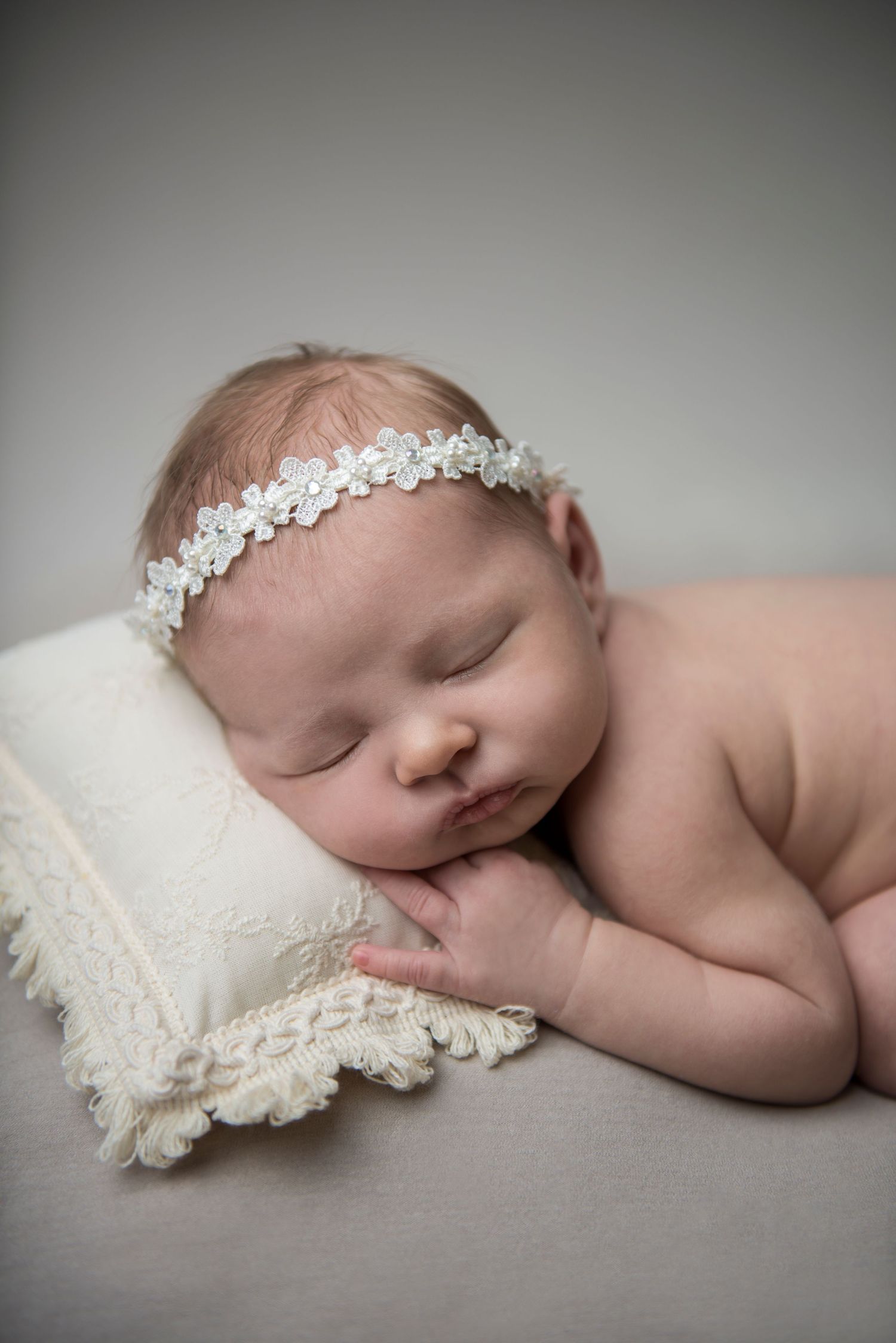 Sleeping baby with lace headband resting on textured white pillow in soft lighting.