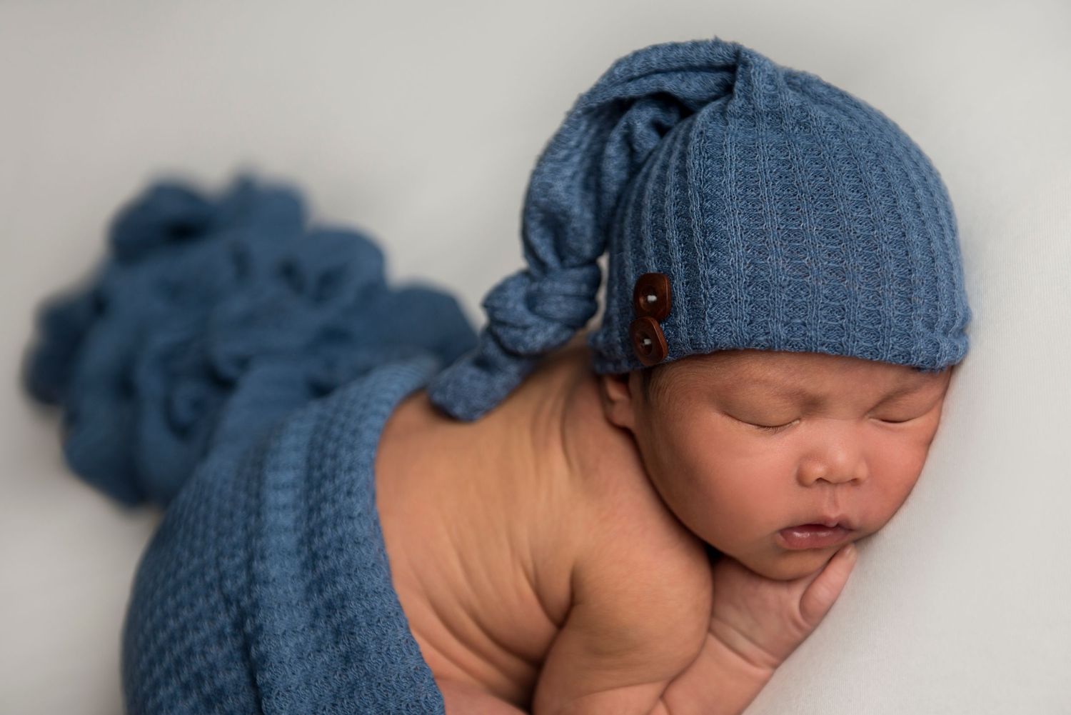 Sleeping newborn wearing blue knit hat and wrap in artistic pose.