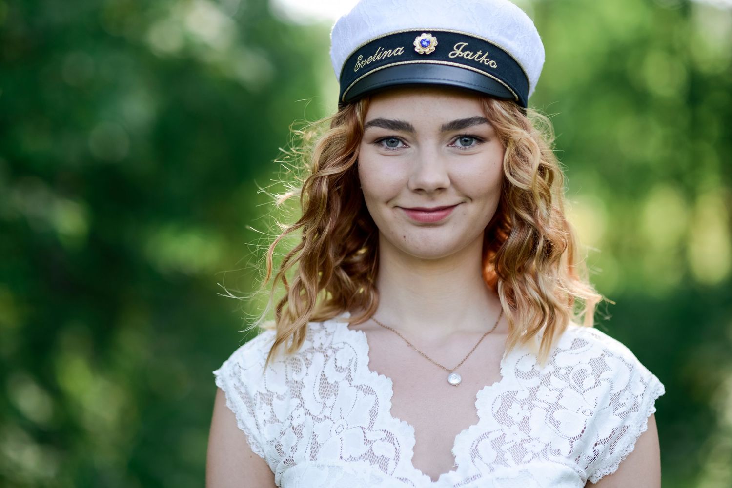 Outdoor portrait in white lace top and sailor cap against green foliage.