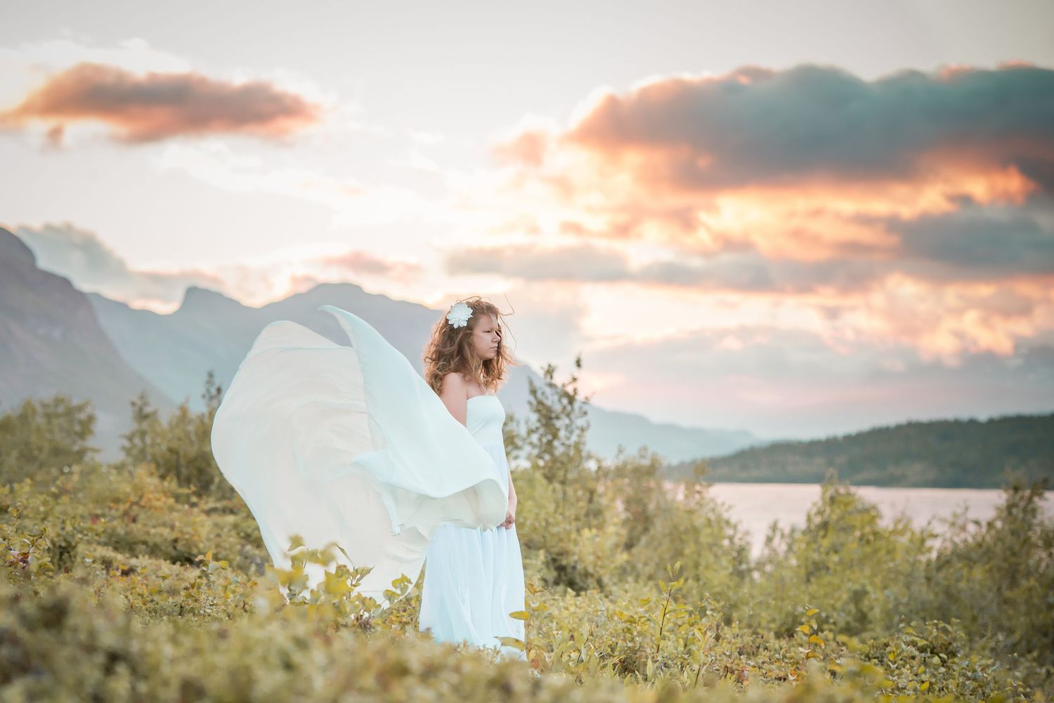 Bride in flowing white dress with floral crown stands in nature at sunset with mountains and lake backdrop.