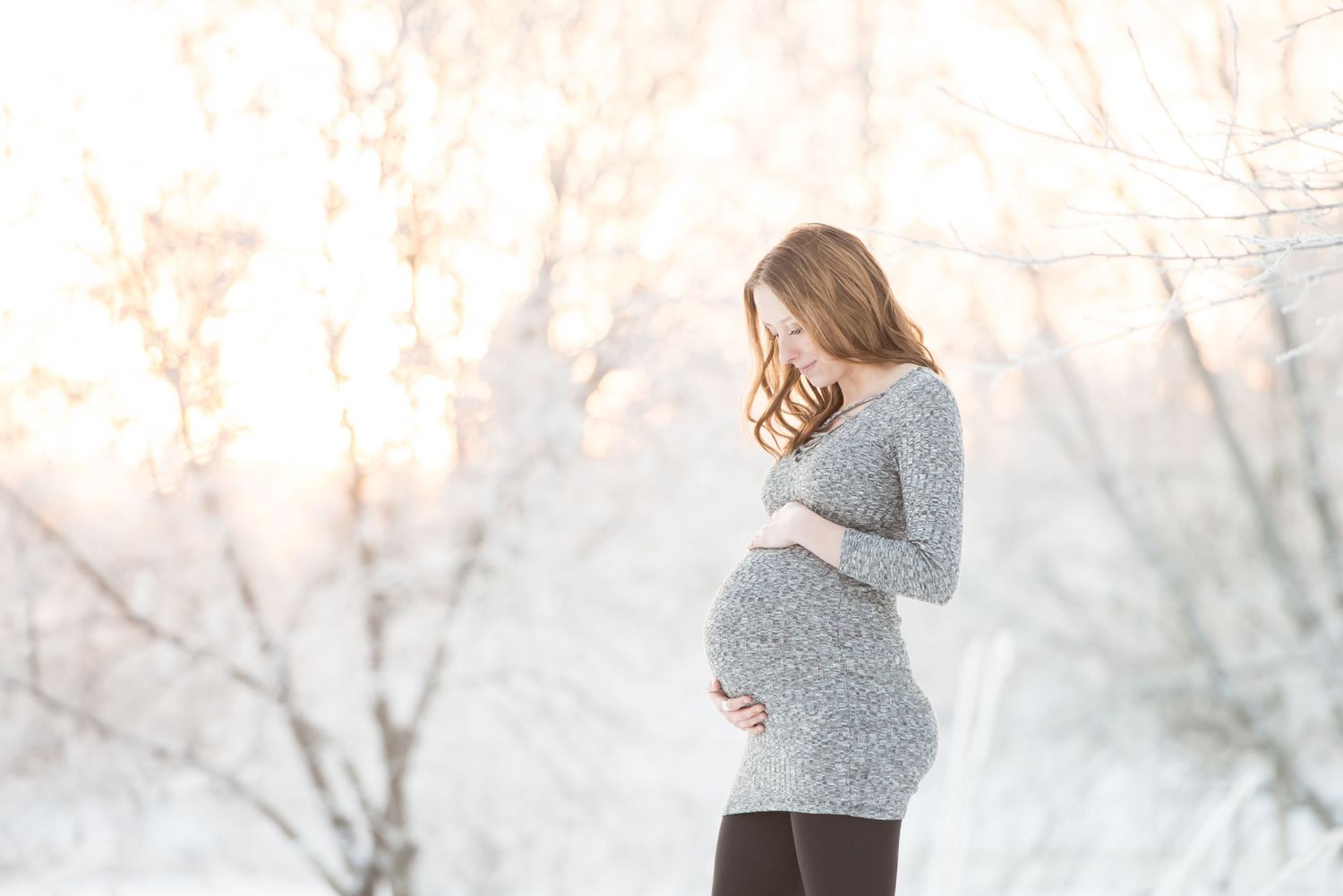 Pregnant woman in gray sweater cradling her baby bump outdoors in a snowy winter landscape.