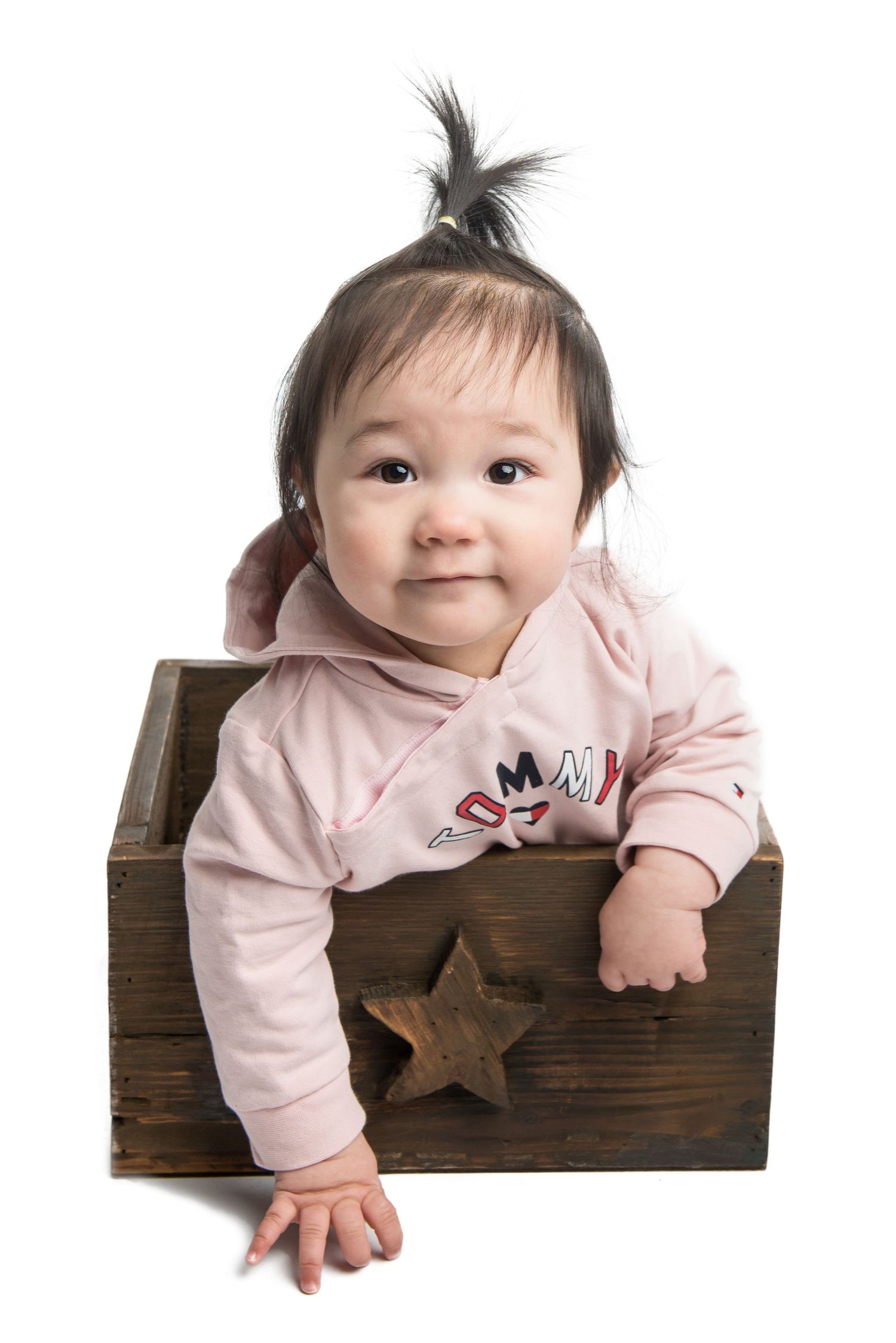 Baby in pink outfit playing in wooden crate against white background.