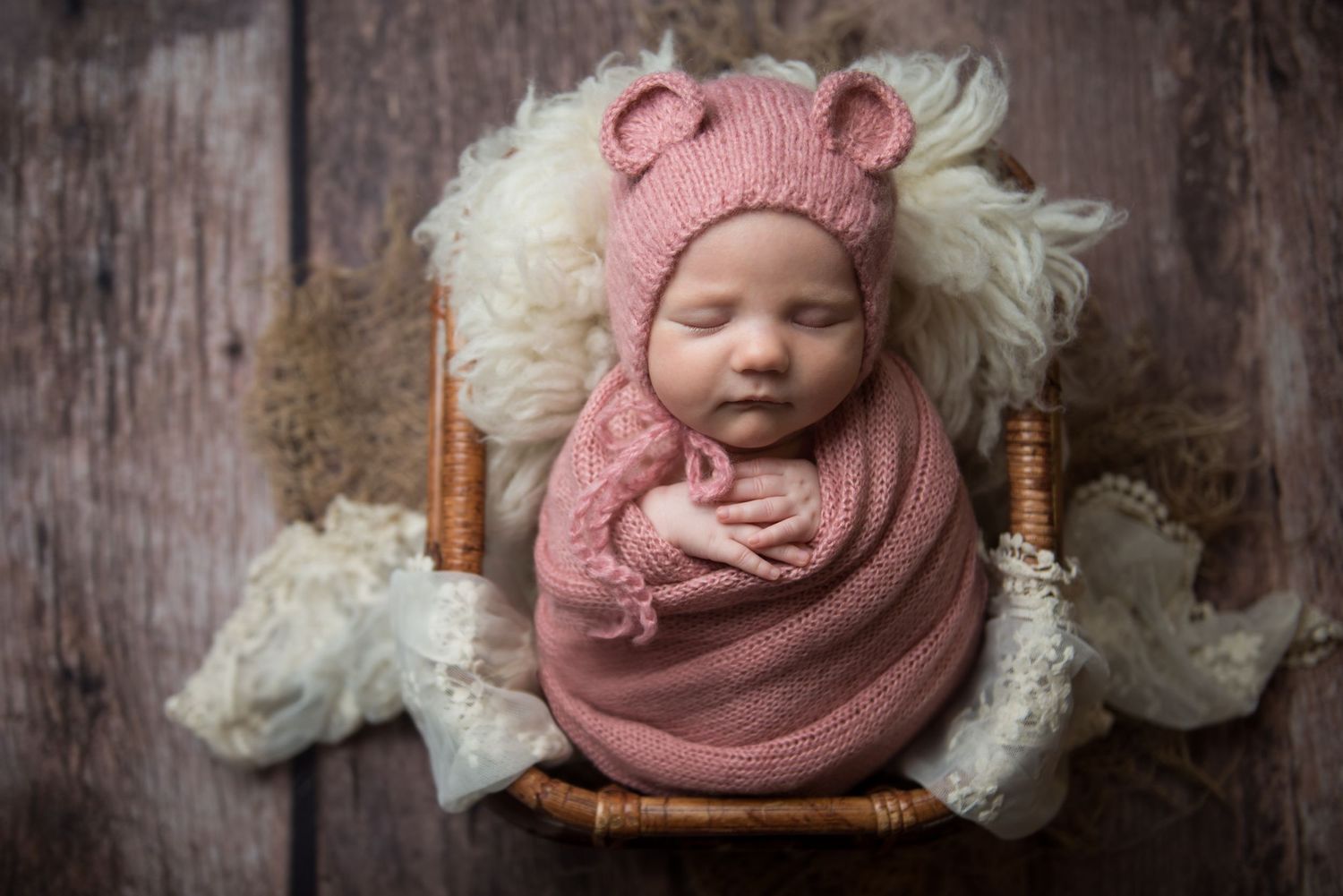 Newborn photography featuring soft pink knit wrap and white fur backdrop against rustic wood paneling.