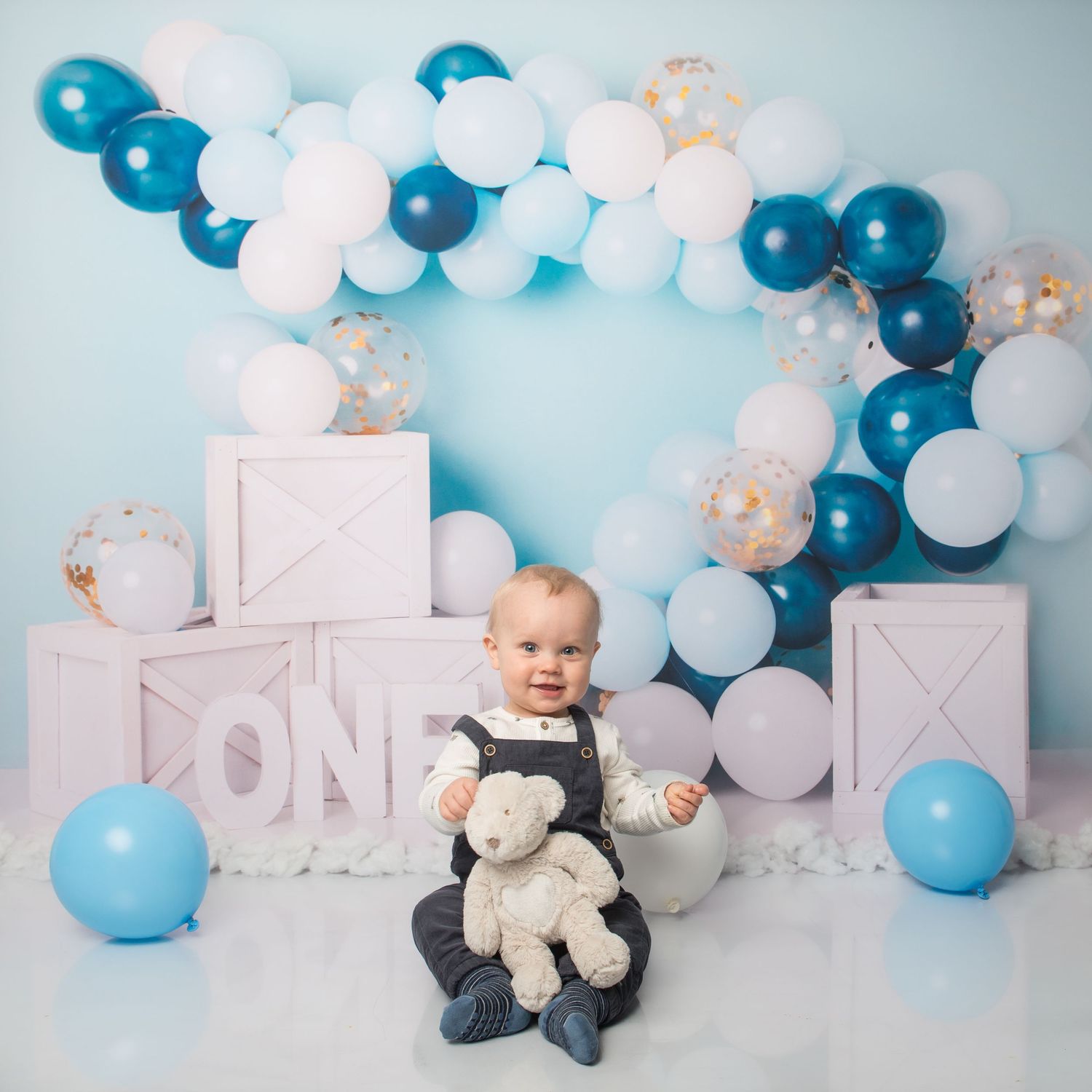 Birthday photo setup with blue and white balloons, decorative blocks, and stuffed animal props.