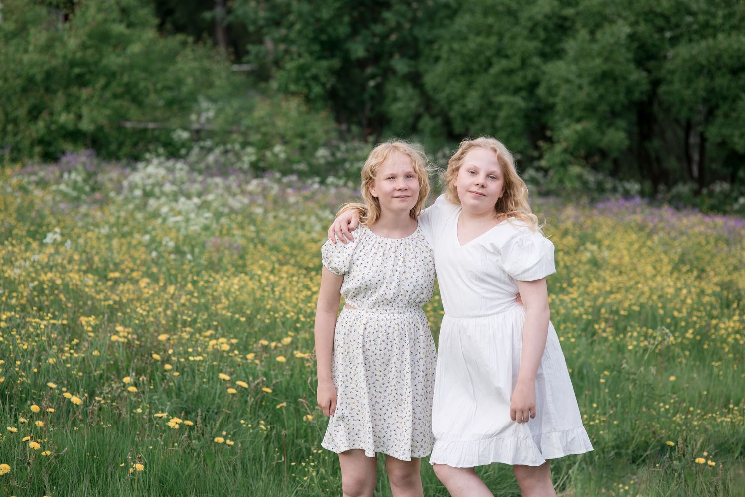 Two blonde women in white dresses pose together in a sunny wildflower meadow with green trees in the background.