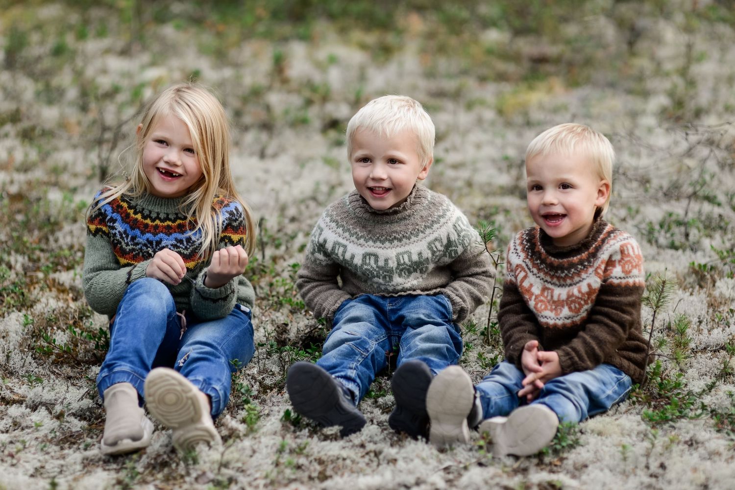 Three young children in matching Nordic sweaters sitting together on frosty ground, smiling happily outdoors.