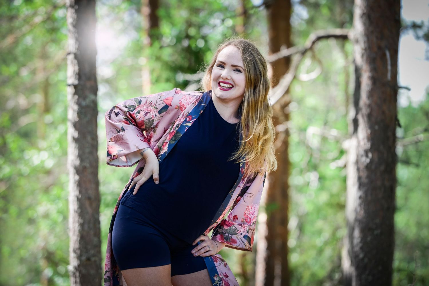 Smiling woman in navy dress and floral kimono posing playfully among tall trees in a bright forest.