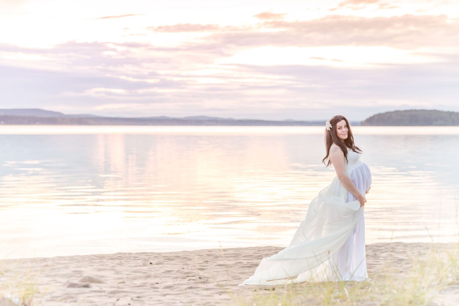 Pregnant woman in white flowing gown standing on sandy beach at sunset with calm water reflections.