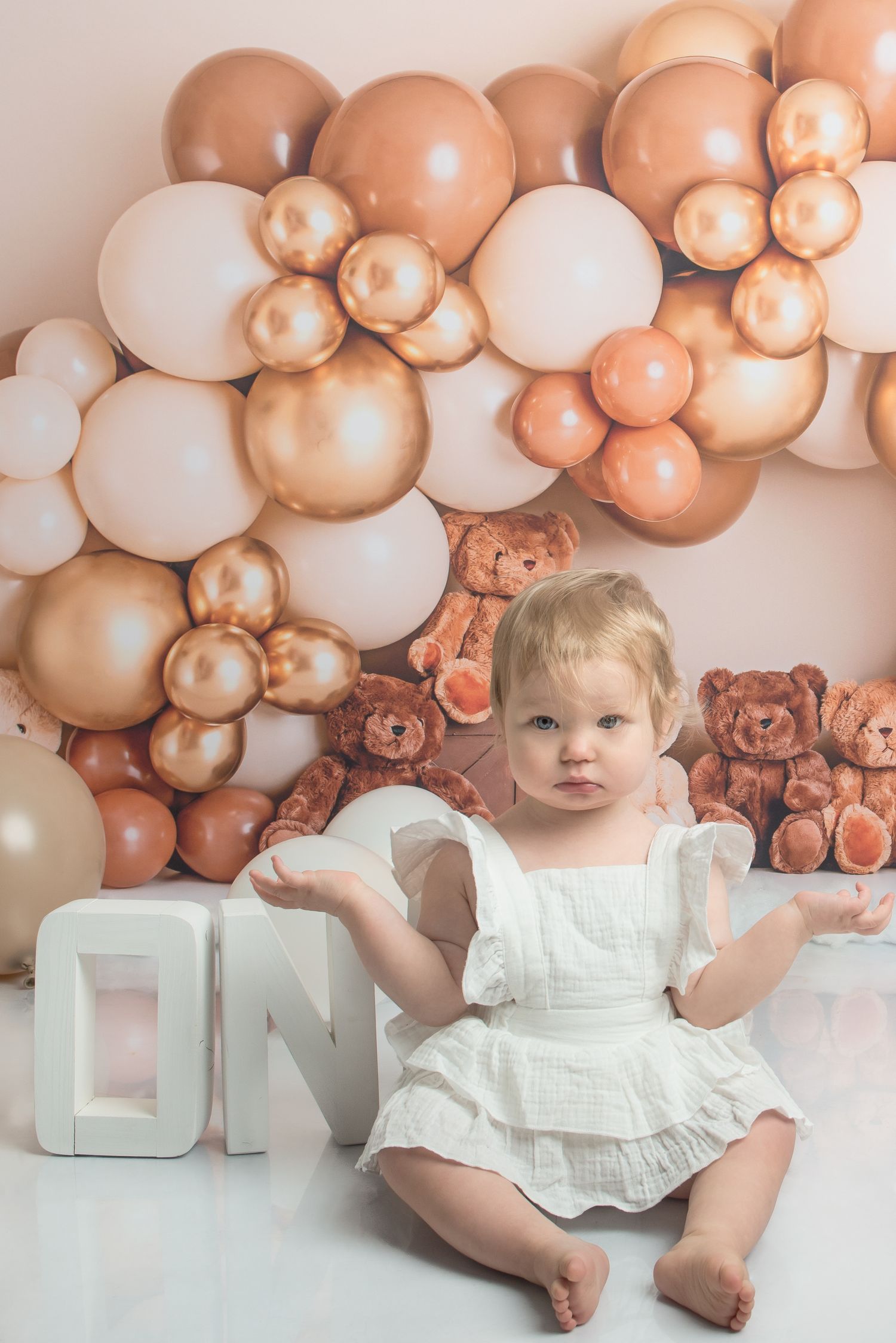 Baby girl in white dress sits before rose gold balloon garland and teddy bears at a first birthday cake smash.