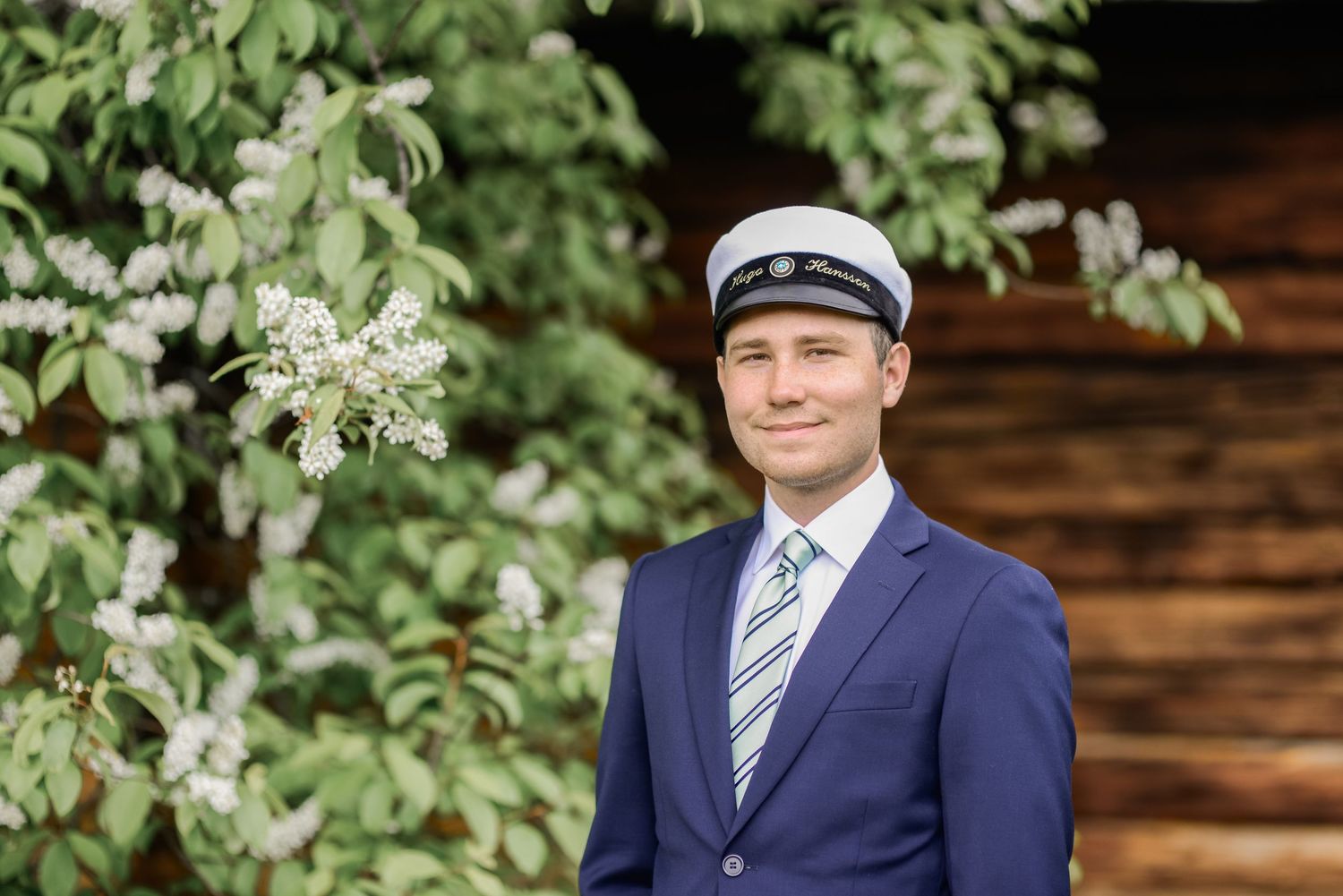 A person in navy suit poses near white flowering trees and rustic wooden wall.