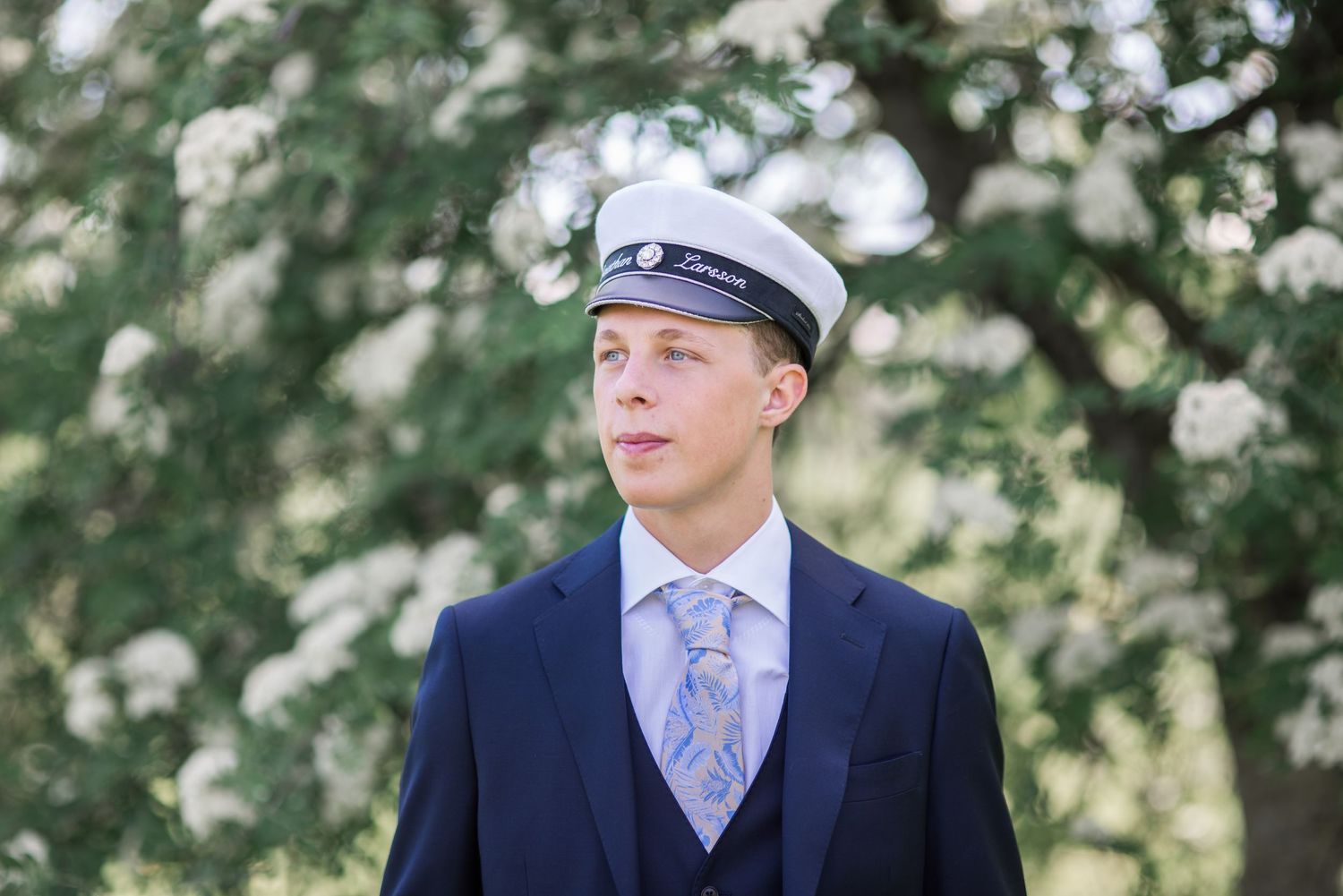 A formal portrait in a navy suit and cap against a blurred floral background.
