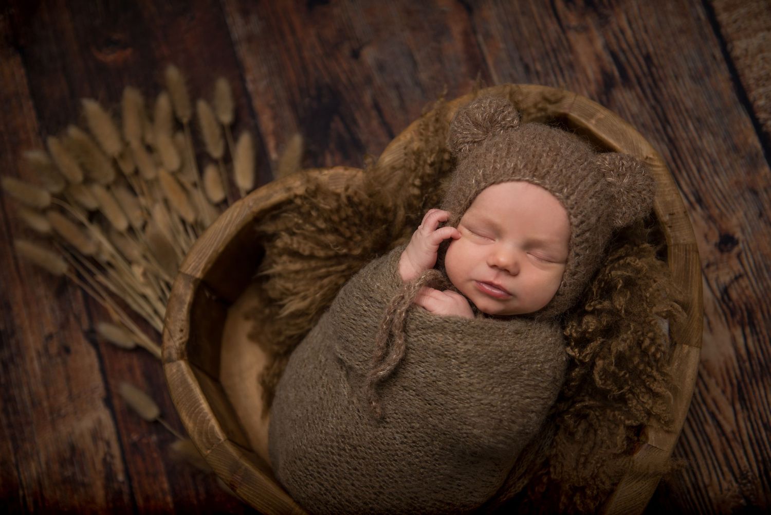 Newborn baby sleeping peacefully on dark wood floor with brown knit wrap.