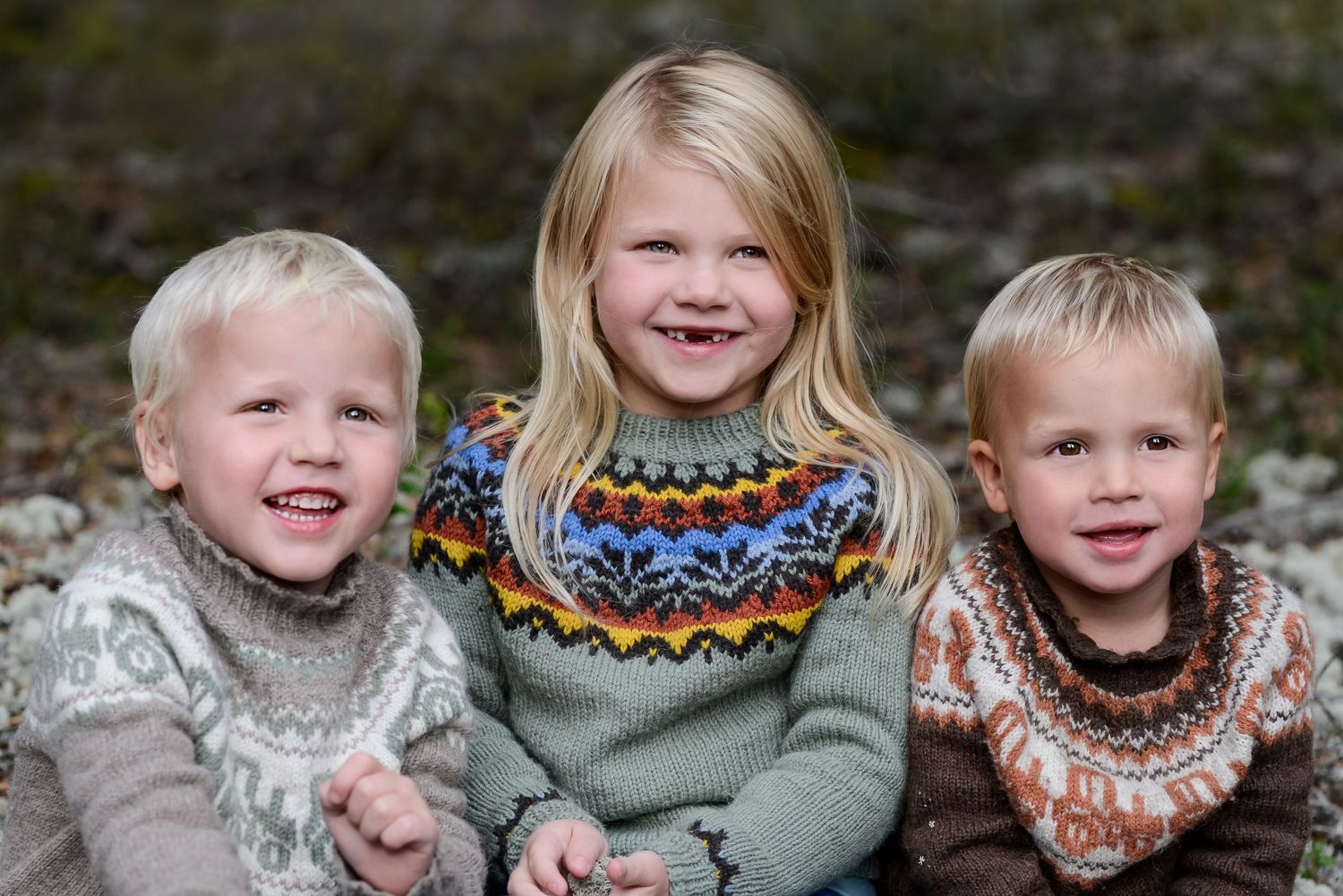 Three children wearing Nordic-style sweaters smile together in outdoor portrait.