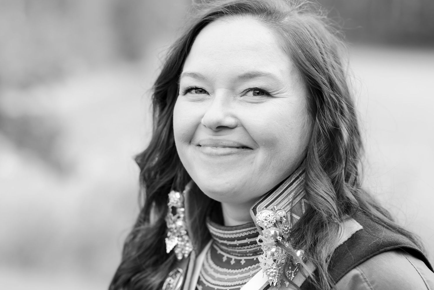 Black and white portrait of a smiling woman with wavy hair wearing traditional Sami cultural attire with decorative jewelry.