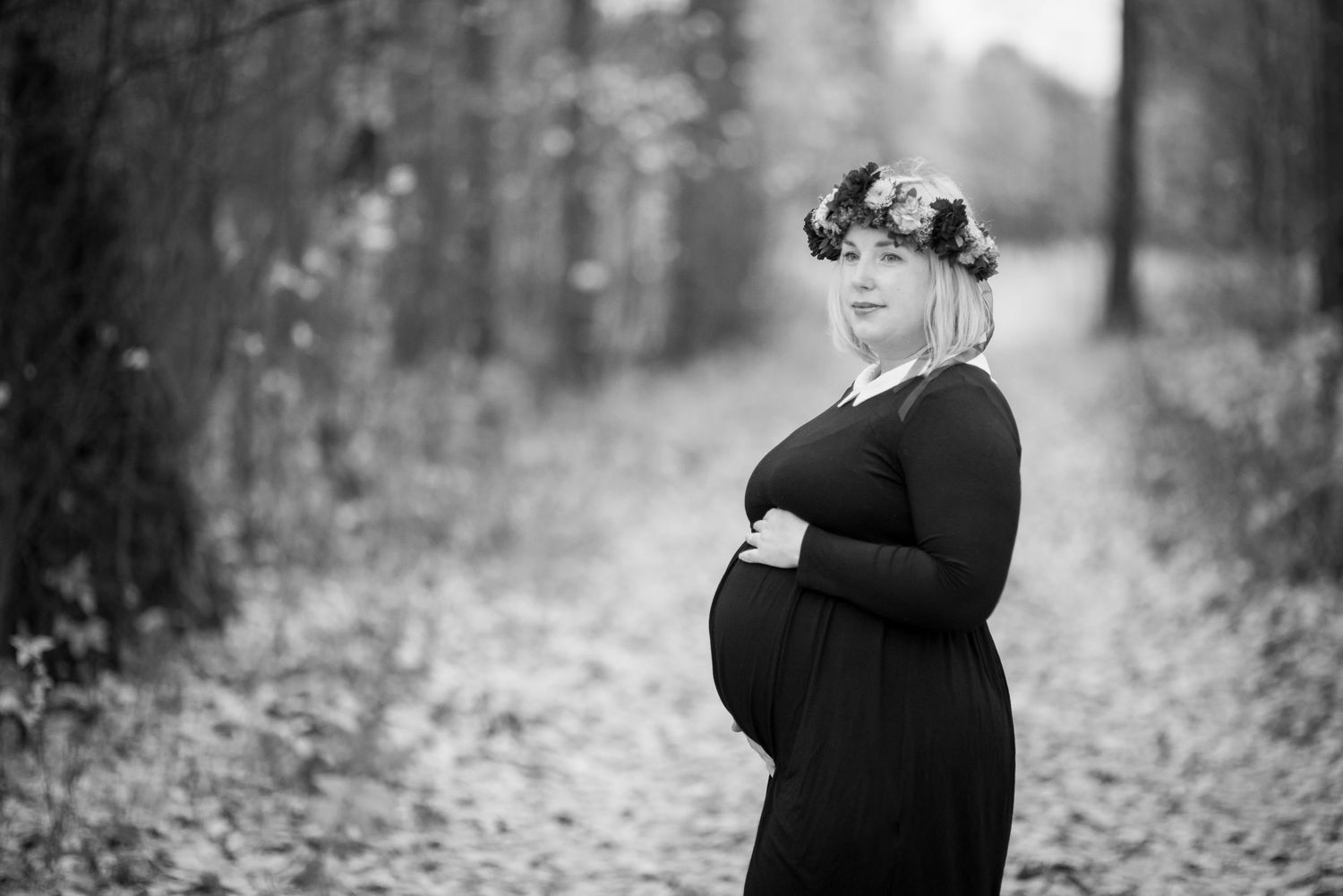 Black and white photo of a pregnant woman in a floral crown and black dress, cradling her belly outdoors.