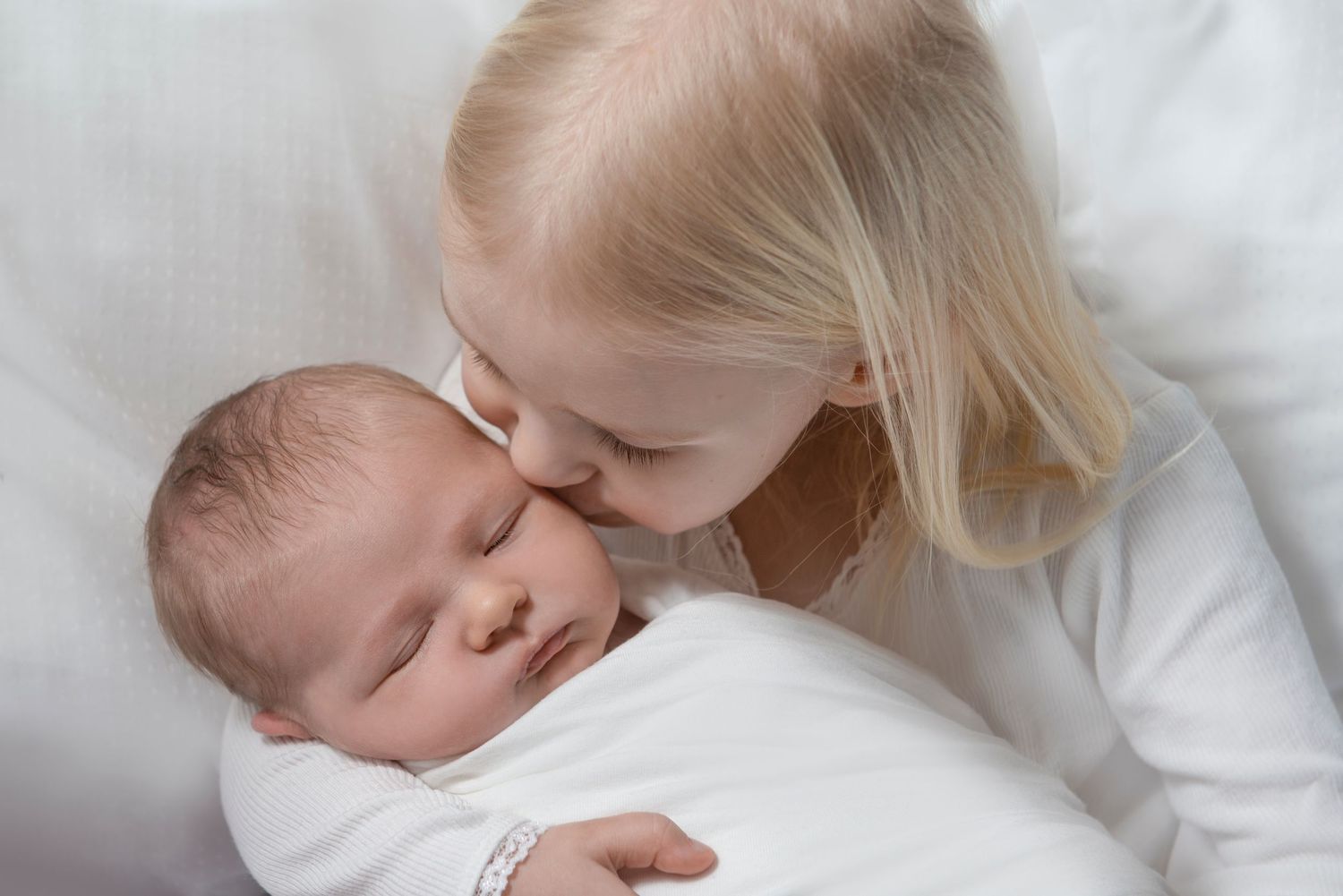 Tender moment between siblings on white bedding in soft natural light.