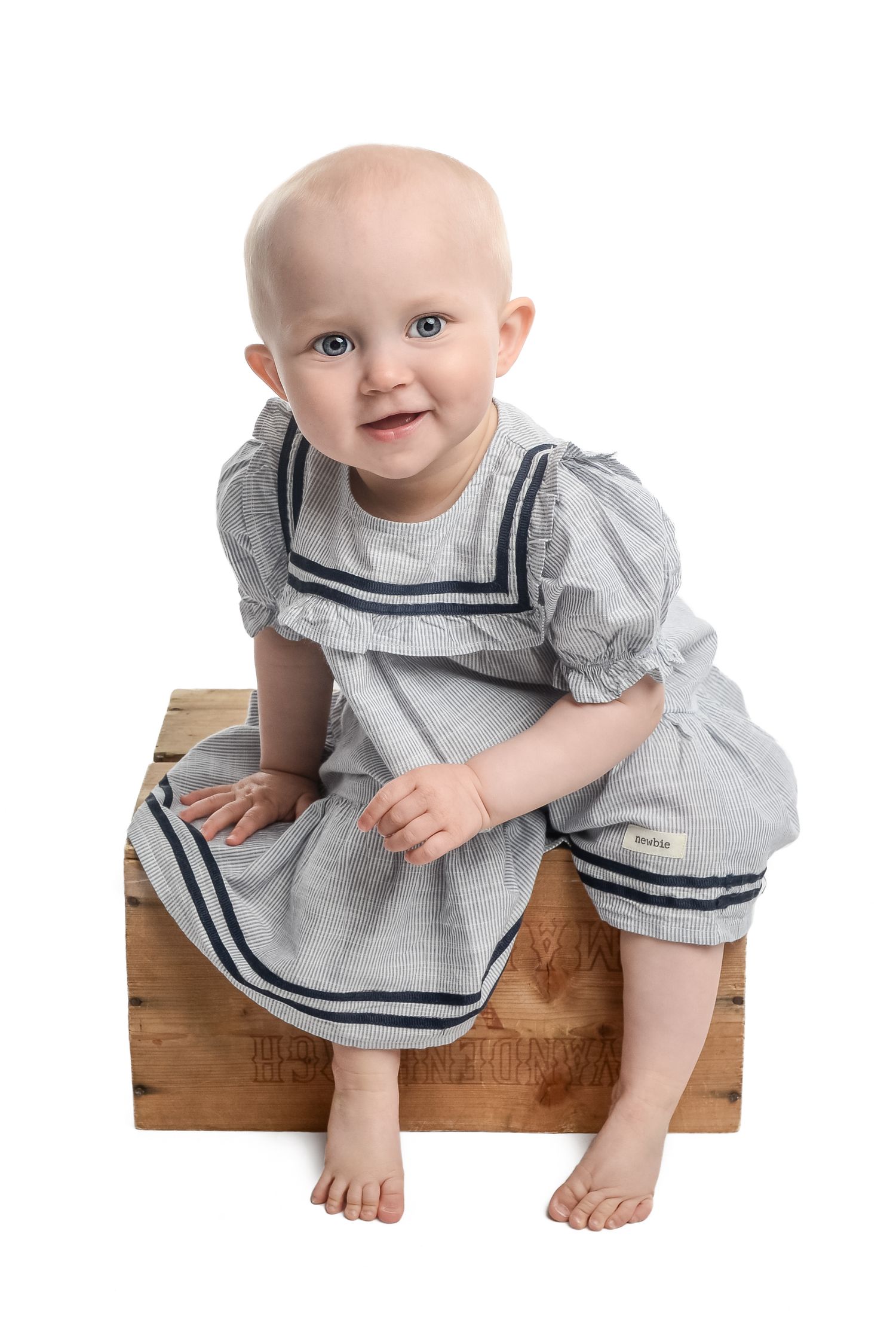 Baby wearing a gray sailor dress with navy trim sits on wooden block showing happy expressions.