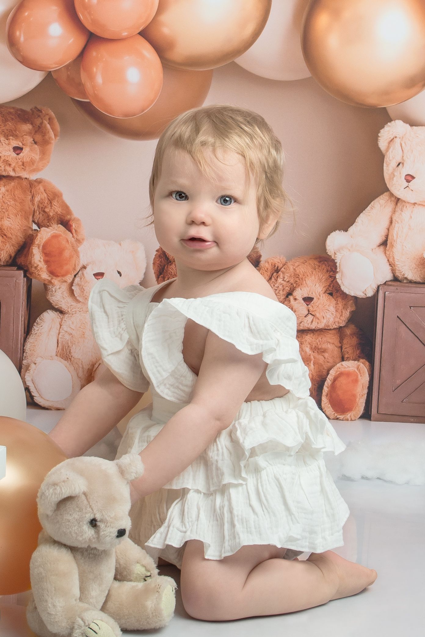 Baby photoshoot scene with pink balloons, plush toys and white ruffled dress against neutral background.