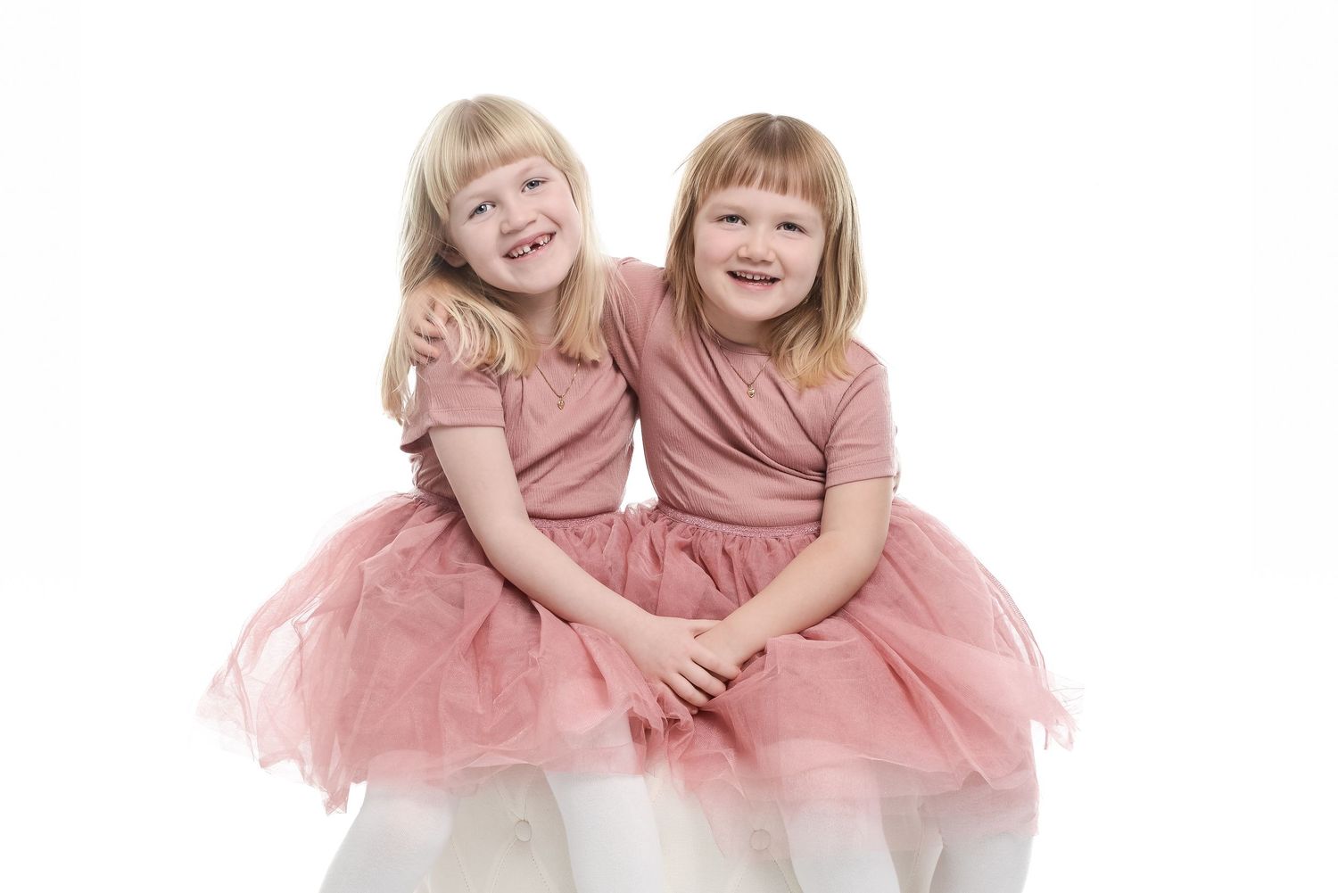 Two children in matching pink dresses embrace while laughing on a white backdrop.