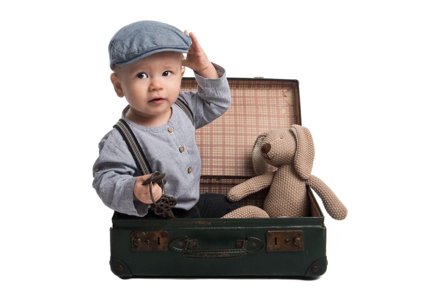 Baby in vintage-style clothing posed with plush toy in antique suitcase.