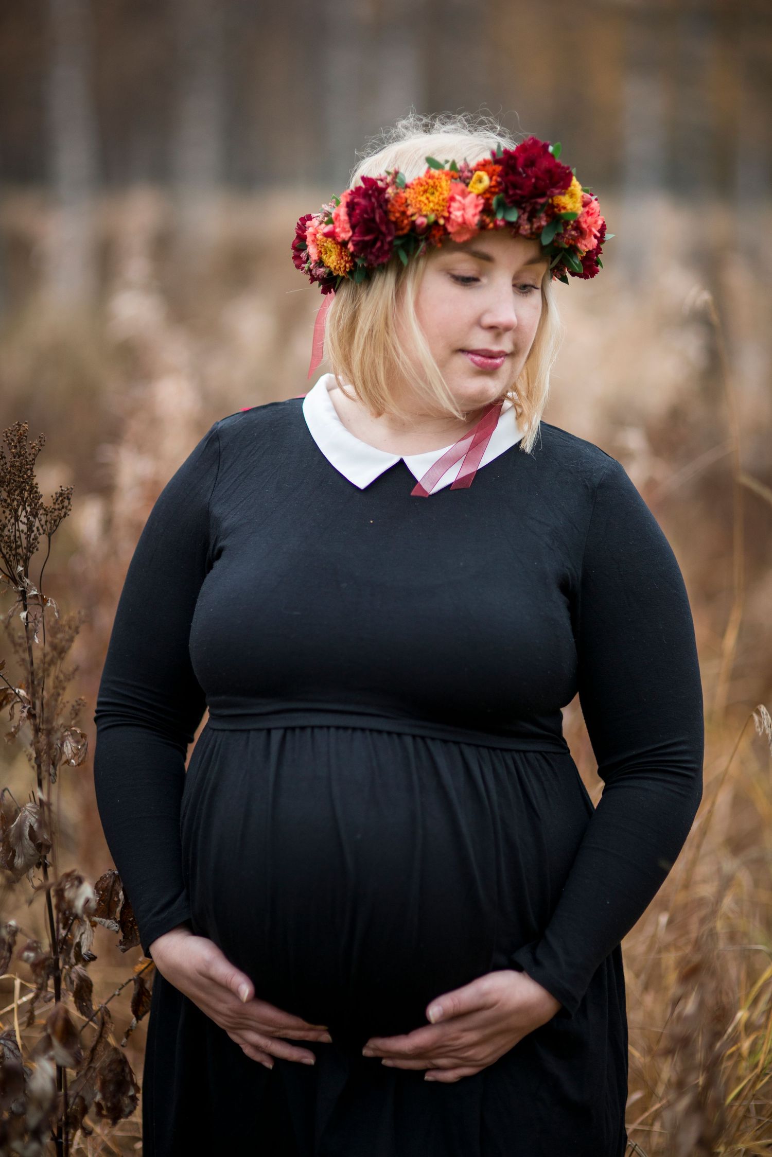 Pregnant woman in black dress and colorful floral crown cradling her baby bump in an autumn field.
