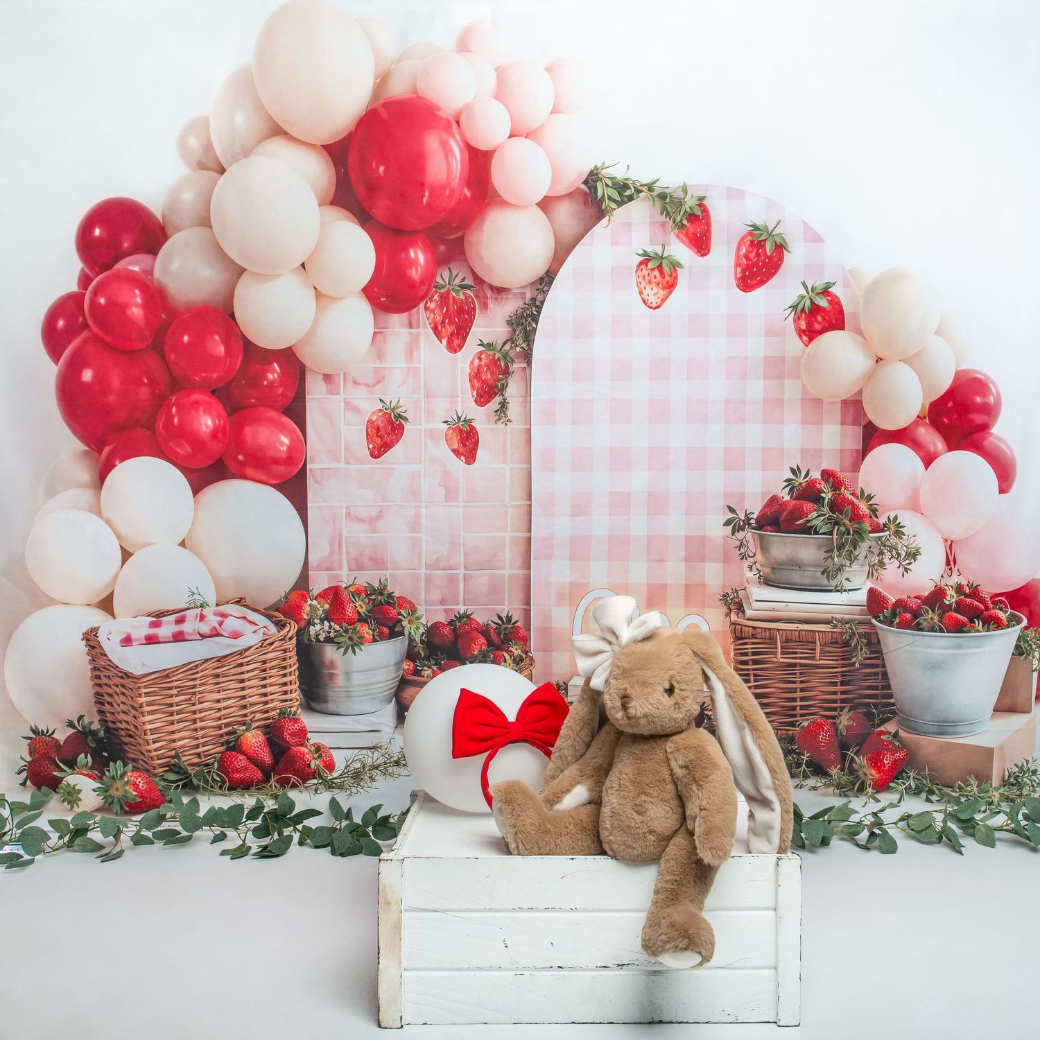 Strawberry-themed party backdrop with pink plaid arch, red and white balloon garland, wicker baskets, and a stuffed bunny with red bow.