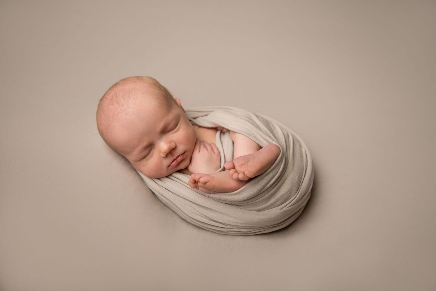 Peaceful newborn baby swaddled in soft beige fabric during studio photography session.