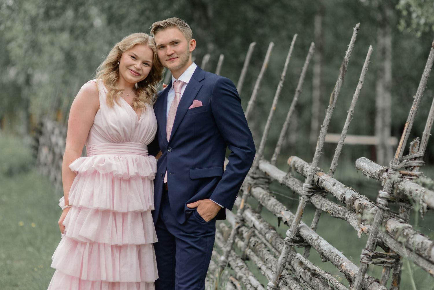 Couple poses by rustic wooden fence during outdoor photo session with natural greenery.
