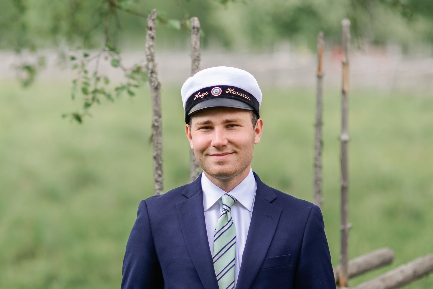 A person in formal attire and baseball cap stands among birch trees in a lush green setting.