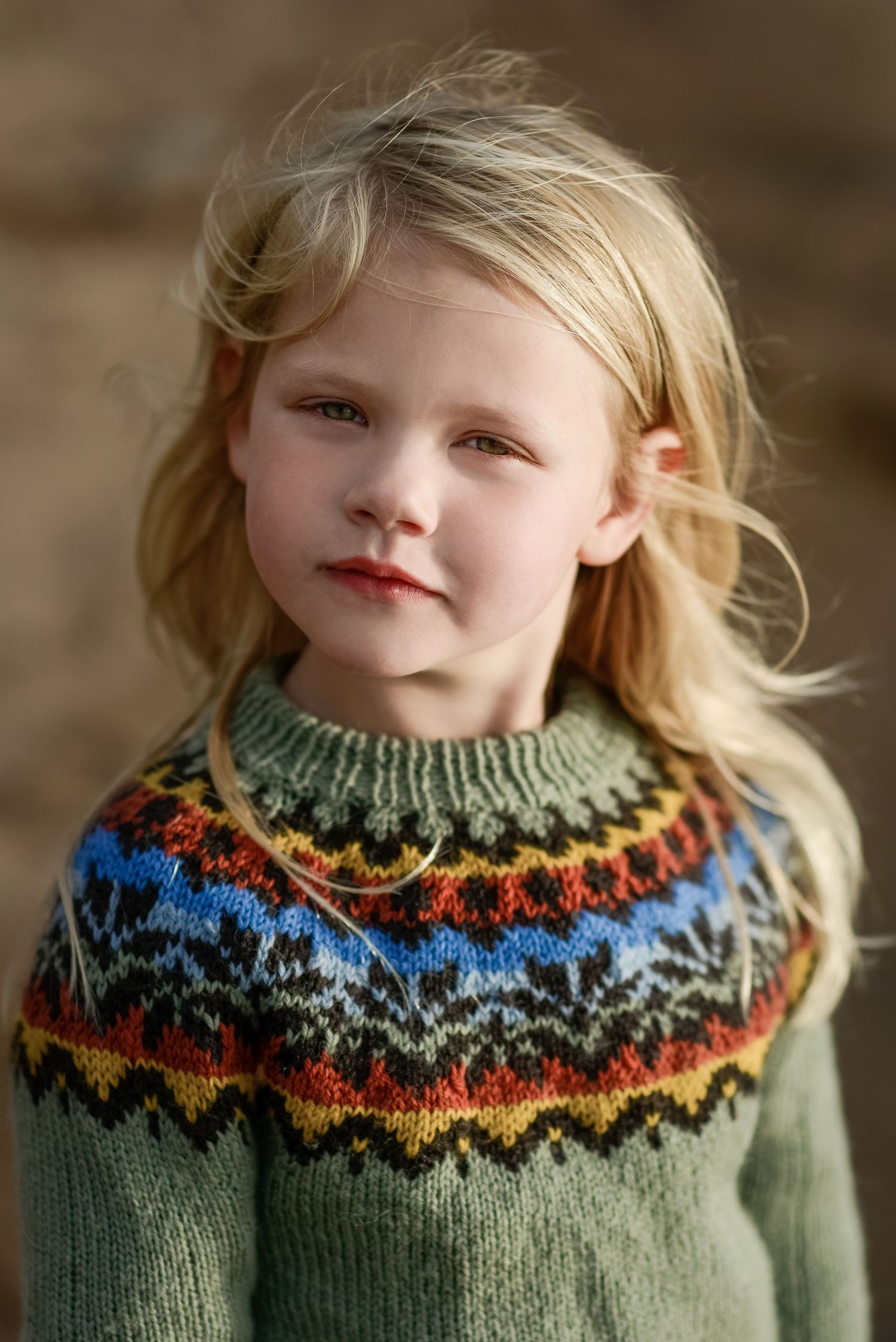Young blonde girl wearing a colorful Fair Isle knit sweater with geometric patterns in green, blue, red, and yellow.